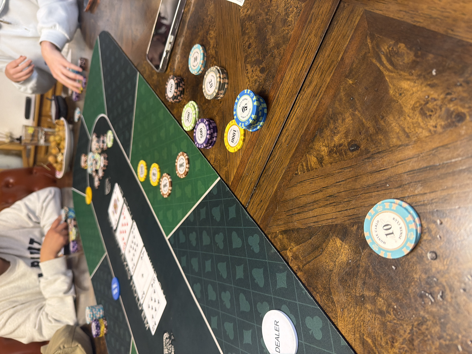 The image captures a close-up view of a poker game in progress, set on a wooden table. The foreground is dominated by a green felt poker mat with a repeating pattern of spades and diamonds. Several stacks of poker chips are scattered across the mat and the table. A single white chip with the word "DEALER" is visible on the left side of the mat. In the midground, a set of playing cards is laid out face down on the mat, with some cards partially revealed. The background shows the arms and torsos of two individuals, also blurred. The person on the left is wearing a white long-sleeved shirt, and their hands are resting on a stack of chips. The person on the right is also wearing a white long-sleeved shirt, and their hands are reaching towards chips. The overall setting suggests a casual game among friends. The image does not provide any specific details or context that would allow for its location to be identified as Xuan Huong - Da Lat, Vietnam.