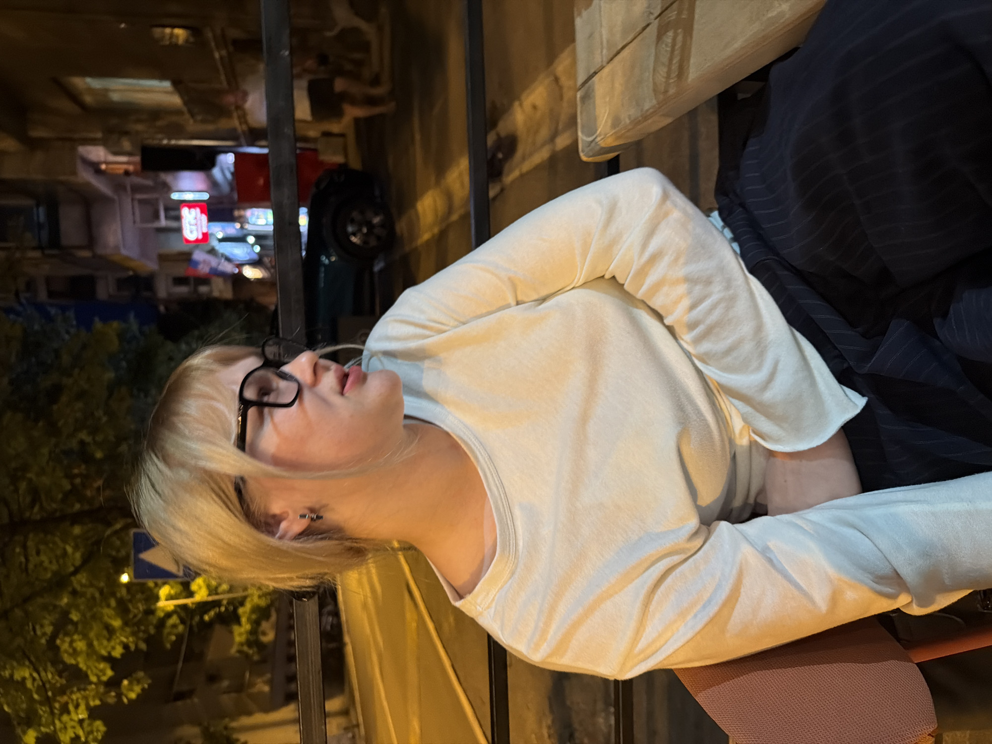The image shows a young Caucasian woman with blonde hair and glasses sitting at an outdoor table at night. She is wearing a white long-sleeved shirt and dark pinstripe pants. The woman is looking to her right, with her arms crossed. In the background, there is a street with cars and buildings. The scene appears to be in the city of Belgrade, Serbia, as indicated by a sign with Cyrillic text and what looks like a Serbian flag in the background. The lighting suggests it is evening or night.