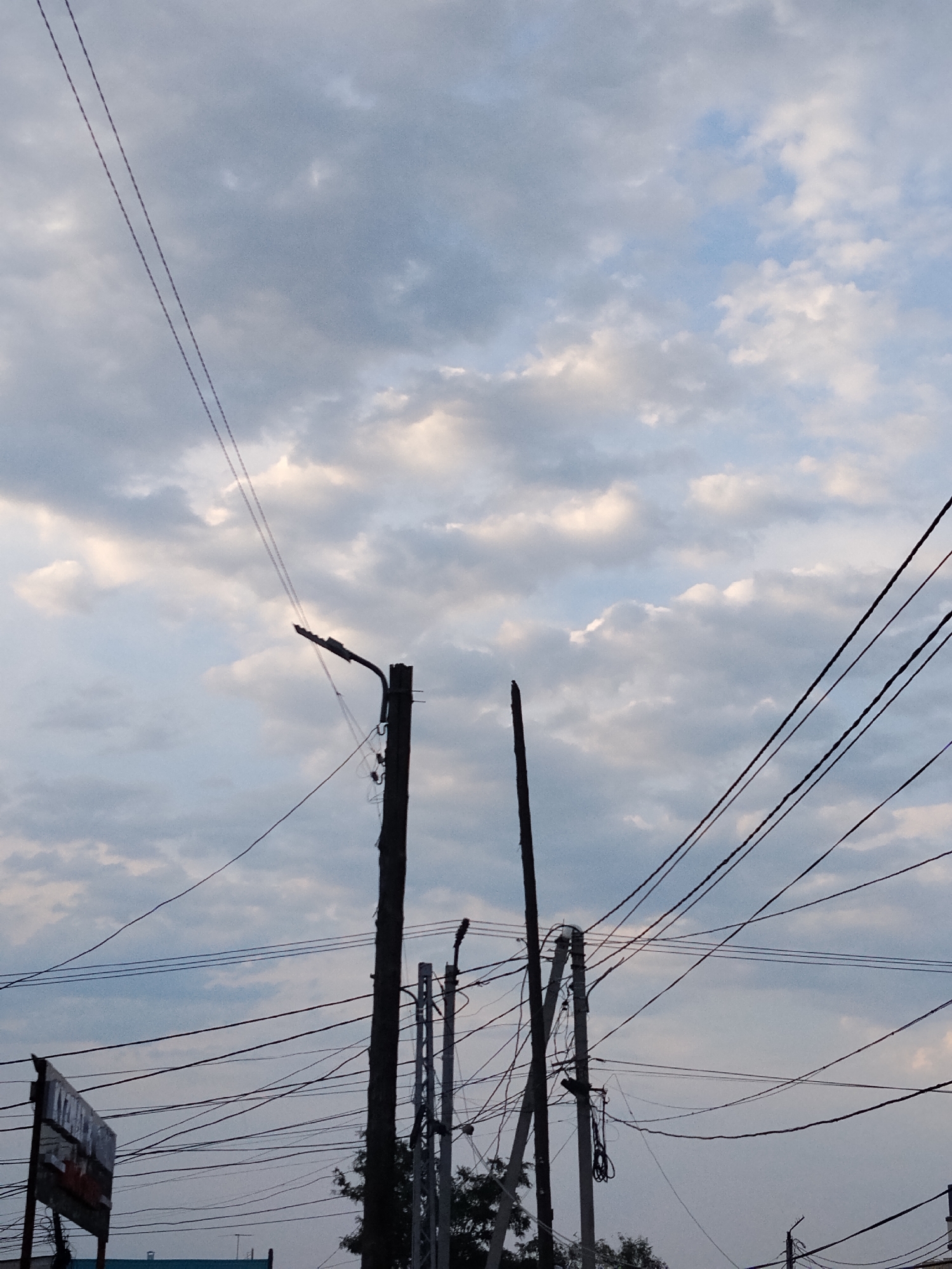 A low-angle shot looks up at a cloudy, overcast sky. Thick, dark power lines crisscross the frame, silhouetted against the lighter sky. Two wooden utility poles are visible, one on the left with a bent, dark lamp attached to it, and another on the right with a broken-off top. A few trees can be seen peeking out from behind the poles and wires, their branches dark against the sky. A signpost with a faded, rectangular sign is partially visible on the left edge of the frame. The time of day appears to be late afternoon or early evening, with the sky a mix of pale blue and grey clouds. The overall mood is somewhat somber and utilitarian, with the focus on the infrastructure of the city against the backdrop of a muted sky. There is no discernible activity taking place.