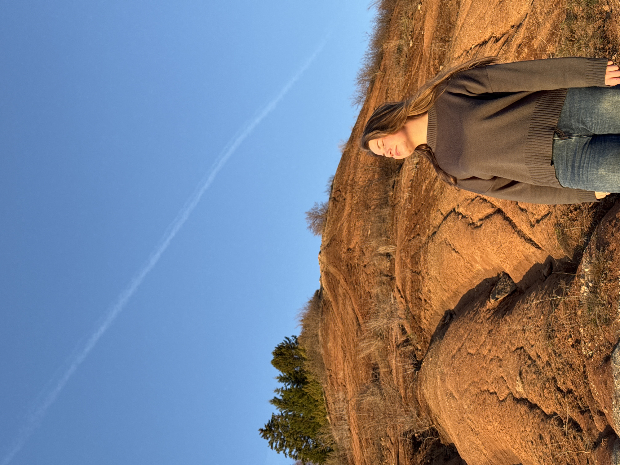 A woman in a gray sweater and blue jeans stands on a reddish-brown, rocky hillside with sparse vegetation. She faces away from the camera, her eyes closed. Above her is a clear blue sky with a contrail. The location is near Ольховка, Russia, based on the reddish rock formations common to that area.