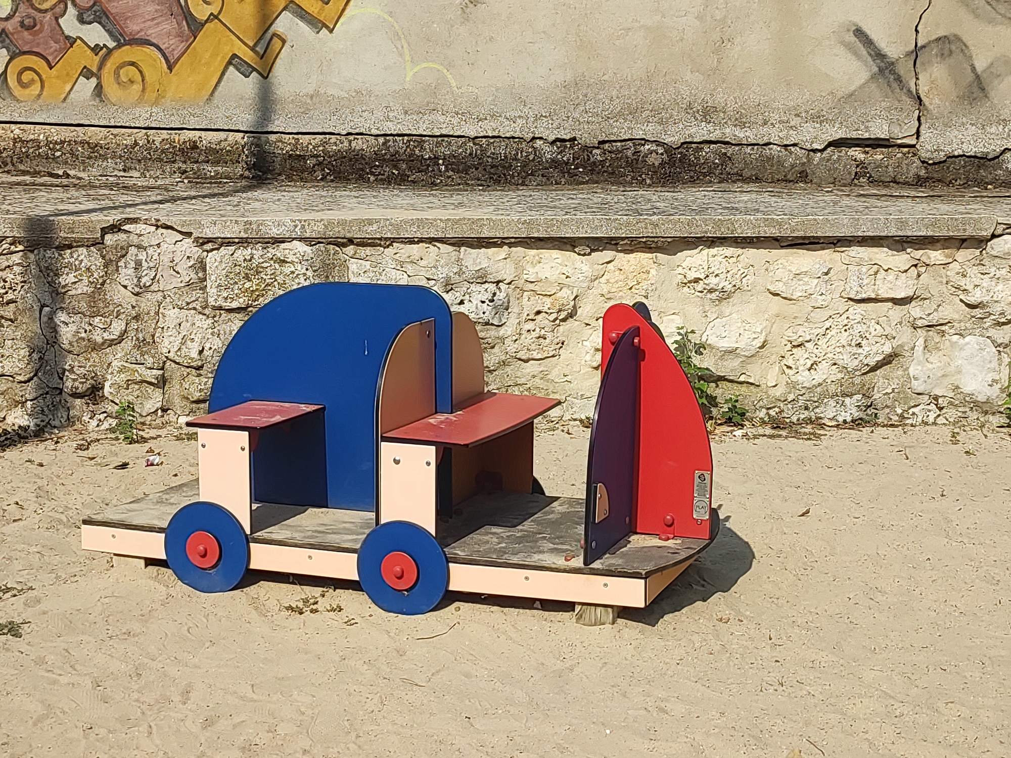 The image shows a children's play structure resembling a train engine and carriage, made of colorful wood and metal, situated on a sandy surface. The structure is primarily blue and red, with beige supports and tan wheels. In the background, there is a textured stone wall, topped with a concrete ledge, and further up, a wall with faded graffiti. The overall environment suggests an outdoor public space, possibly a park or playground, in the city of Traspinedo, Spain. There are no people visible in the image. The lighting indicates it is daytime.