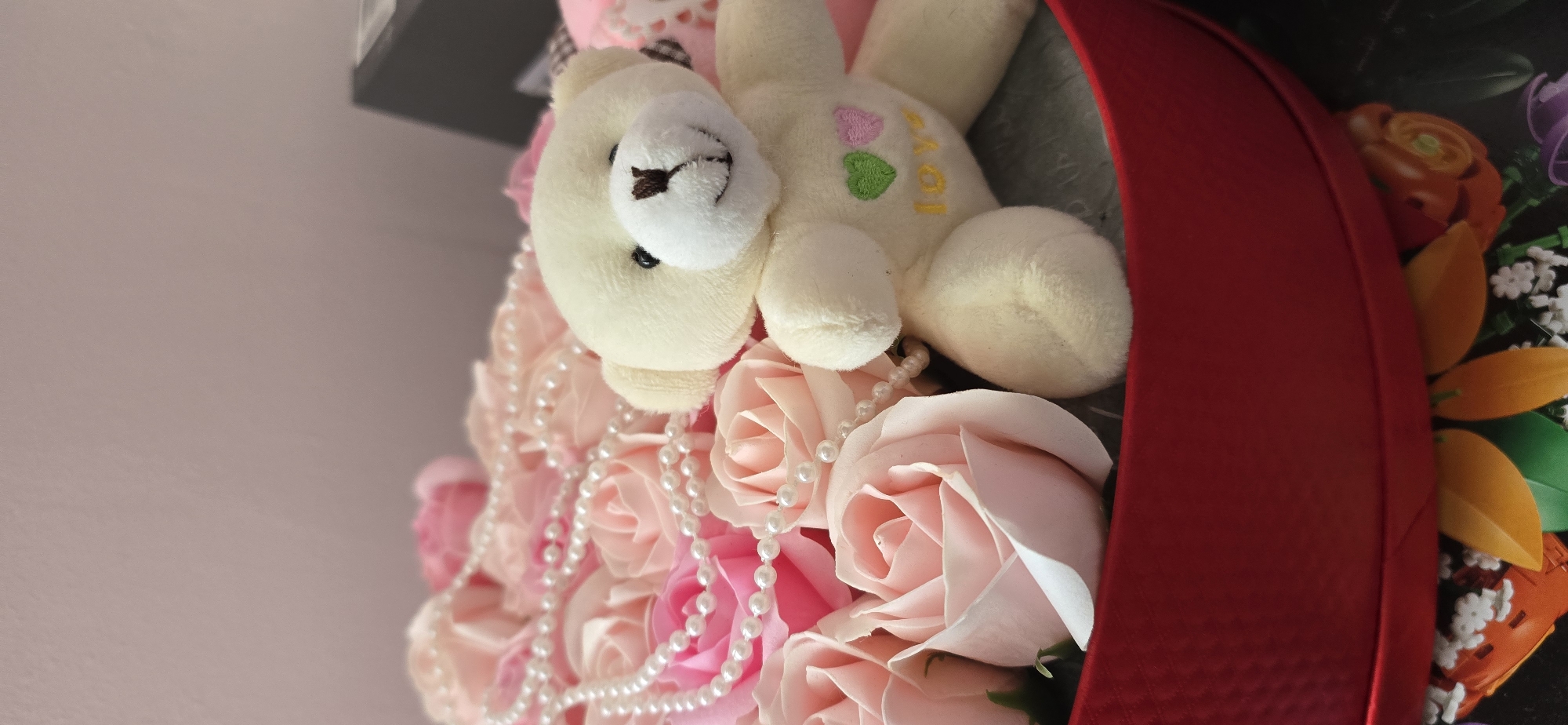 This image displays a close-up of a decorative arrangement, possibly a gift bouquet or box, containing artificial flowers and a plush toy. The primary subjects are a small, cream-colored teddy bear and numerous pink and peach-colored artificial roses.
The teddy bear, positioned centrally towards the upper part of the frame, is lying down with its head slightly raised. It has black bead eyes and a brown stitched nose and mouth. On its torso, the word "LOVE" is embroidered in yellow, accompanied by a pink heart and a green heart.
Cascading over and around the artificial roses are several strands of white faux pearls, adding a touch of elegance. The roses, appearing to be crafted from soap or silk, are arranged densely, filling the lower and central parts of the image with varying shades of pink and peach. A vibrant red ribbon or fabric trim outlines the right side of the arrangement, suggesting it is contained within a box or wrapped.
In the background on the far right, partially obscured by the red trim, are glimpses of other artificial floral elements in orange, green, and white. The background on the left consists of a plain, light-colored surface. The overall impression is one of a thoughtful and romantic gift. The original image is oriented sideways, appearing as if rotated 90 degrees clockwise from its natural upright position.