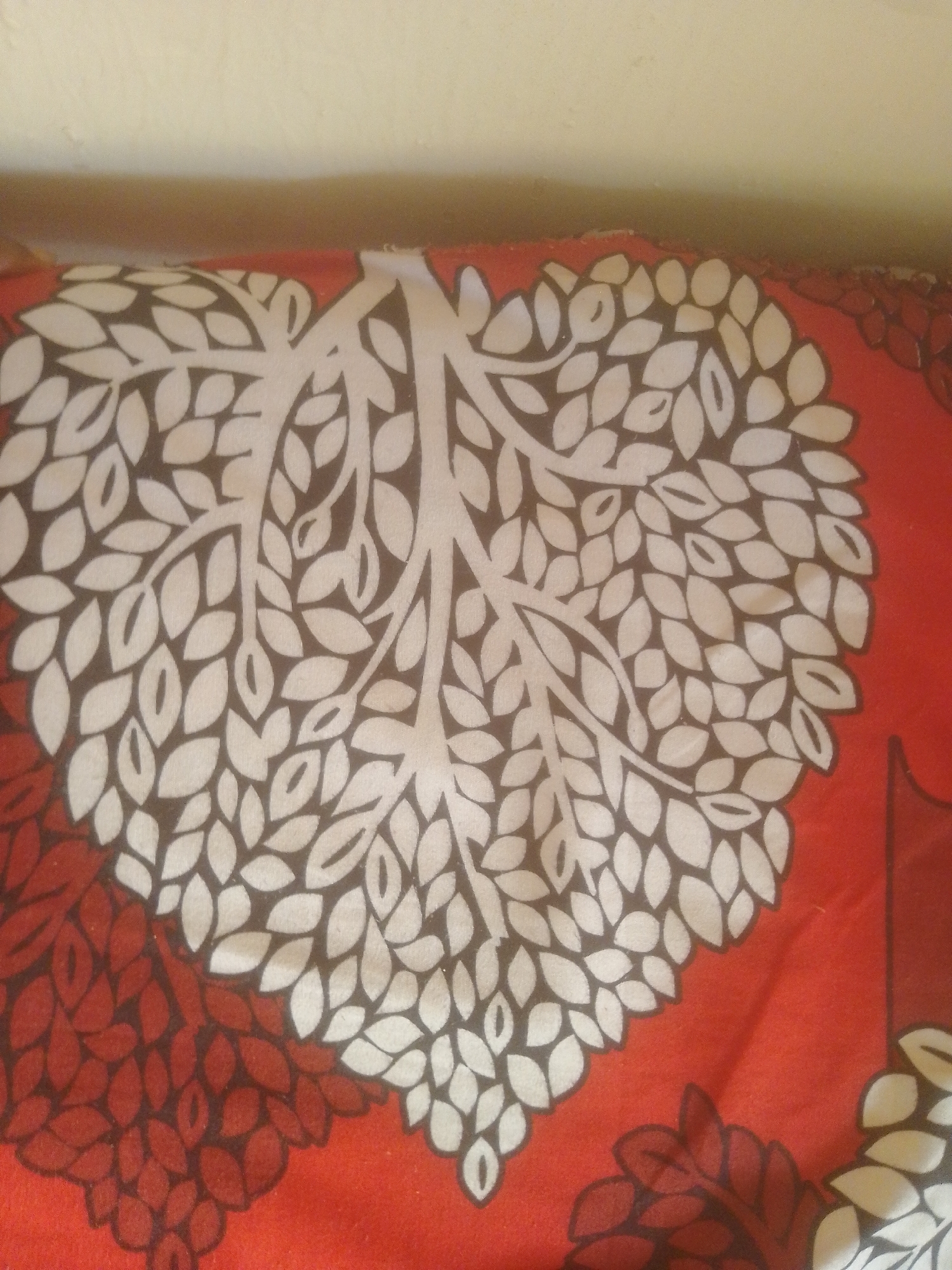 The image shows a close-up view of a red patterned fabric, likely a pillowcase or bedsheet, featuring a stylized tree with heart-shaped leaves. The tree's branches and some leaves are depicted in white and brown, creating a striking contrast against the red background. The fabric appears to be lying on a surface, with a wall visible in the upper portion of the image. The location is identified as Majemu, Nigeria, but no specific urban context is discernible from the image itself.