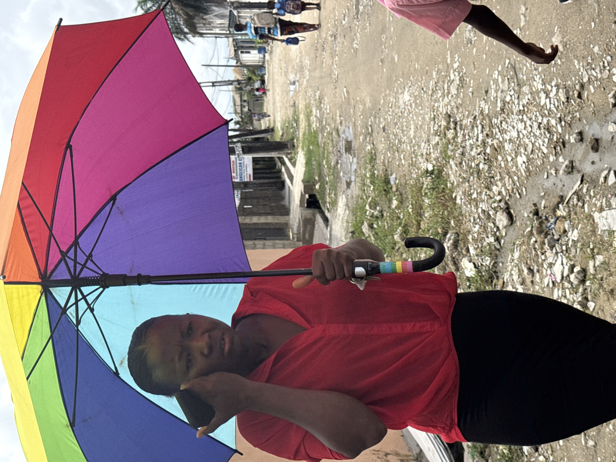 This portrait-oriented image, rotated 90 degrees clockwise, captures an outdoor scene in Ado, Nigeria, likely during the daytime under bright, possibly hazy, conditions. The main subject is a person, seemingly female, with dark skin and short dark hair, wearing a red short-sleeved shirt and black pants. She is holding a large, open rainbow-colored umbrella (displaying panels of red, orange, yellow, green, blue, and purple) with her right hand on the handle. Her left hand is resting near her face, and she looks directly at the camera with a neutral, slightly contemplative expression.
The background features a dusty, unpaved road or pathway strewn with small rocks and debris, with scattered patches of dry grass. Some areas of the ground appear damp or darker. Further down the path, several blurry figures are visible walking, one carrying what looks like a container. In the upper right corner, the lower leg and a pink garment of another person are partially visible. Beyond the road, simple, low-rise buildings and structures can be discerned, indicating a residential or community area. A white sign in the background, partially obscured by the umbrella, shows visible text including "O", "ANGLICAN C _ _ _", and "COMMUNITY", suggesting it belongs to an Anglican Church Community. The presence of the umbrella implies a need for protection from sun or rain, commonly experienced in tropical climates.