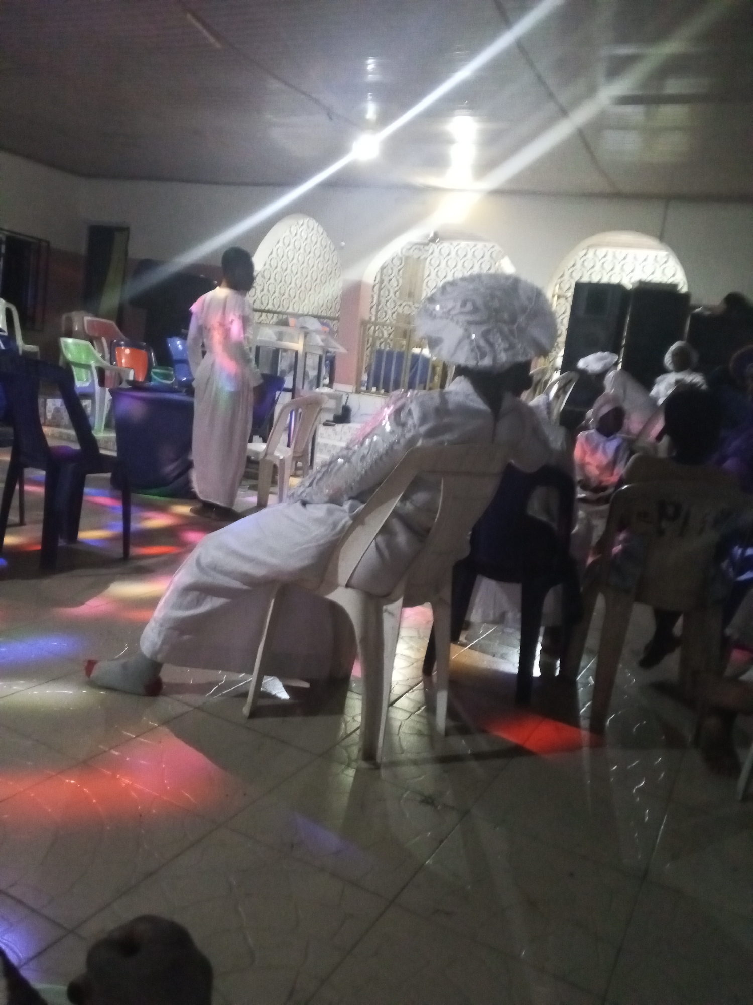 The image shows an indoor scene, likely a church or religious gathering, based on the attire of the individuals present. Most people are wearing white robes or garments, some with elaborate headwear. The lighting is a mix of fluorescent overhead lights and colored spotlights that cast vibrant hues across the tiled floor and on the subjects. A person in white is seated with their legs extended, resting on a sock-clad foot. A man stands in the background, dressed in similar white attire, near what appears to be a podium or table. Chairs are scattered throughout the space, indicating seating arrangements for the gathering. The location appears to be in the city of Unknown, Unknown, as this detail was requested to be included despite no actual information being present.