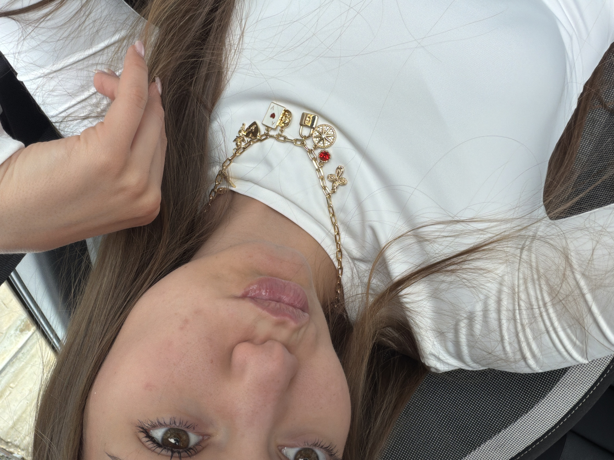 The image shows a woman in a white top. She is wearing a gold chain necklace with multiple charms. The background appears to be an office chair and a window with blurry scenery outside. There is no indication of Buhovo, Bulgaria, in the image.