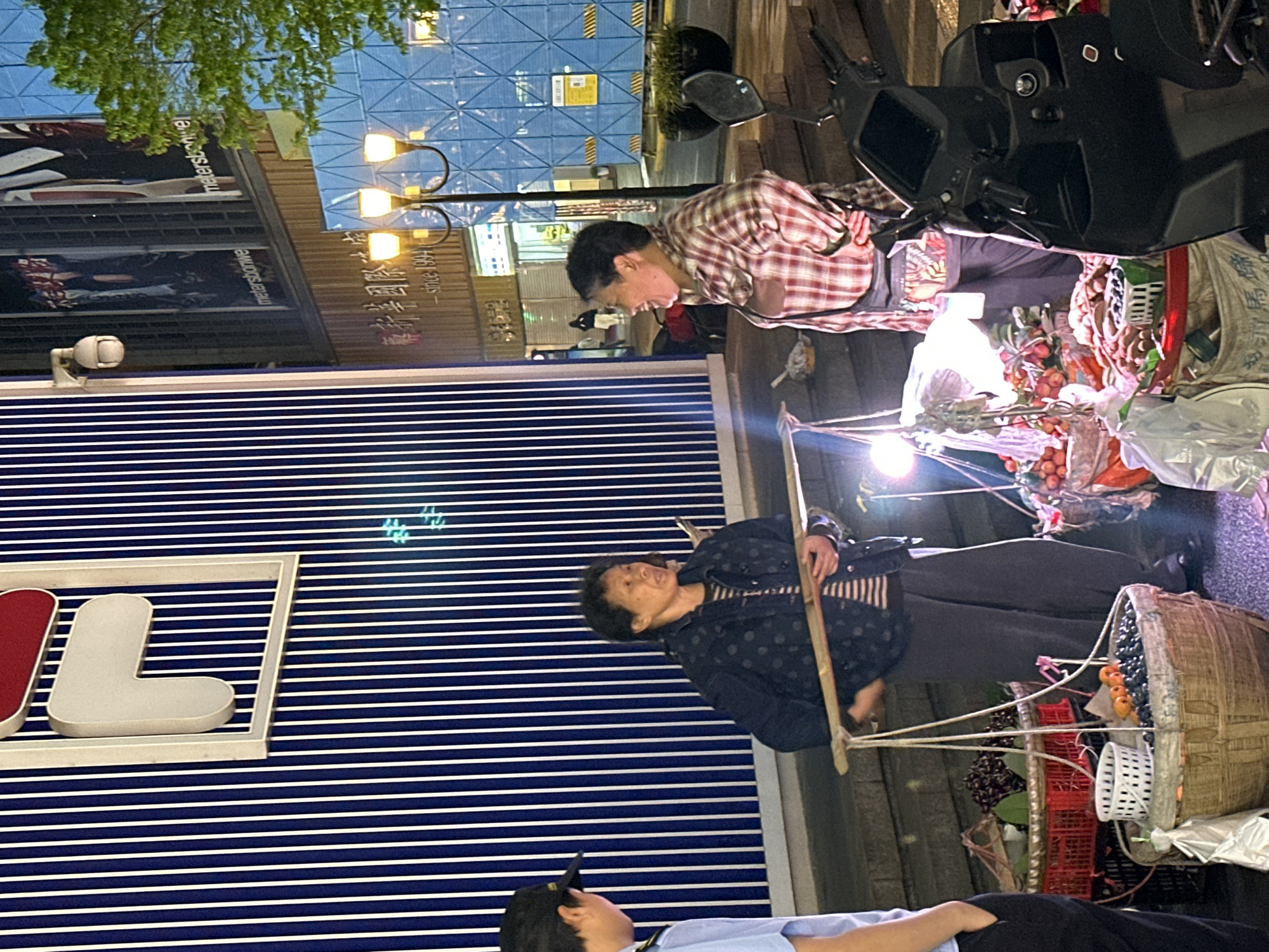 The image captures a street scene in the Yuzhong District of China, likely at night given the artificial lighting. Two elderly vendors are present, selling goods on the sidewalk. One vendor, dressed in a plaid shirt, stands near a motorcycle laden with produce. The other, wearing a polka-dot jacket, is seen with a traditional carrying pole and baskets filled with items, possibly fruits. A police officer in uniform is also visible, seemingly interacting with one of the vendors. The backdrop features a modern building with a blue and white striped facade and visible signage, suggesting a commercial area. Streetlights and a reflective building further contribute to the urban environment.
