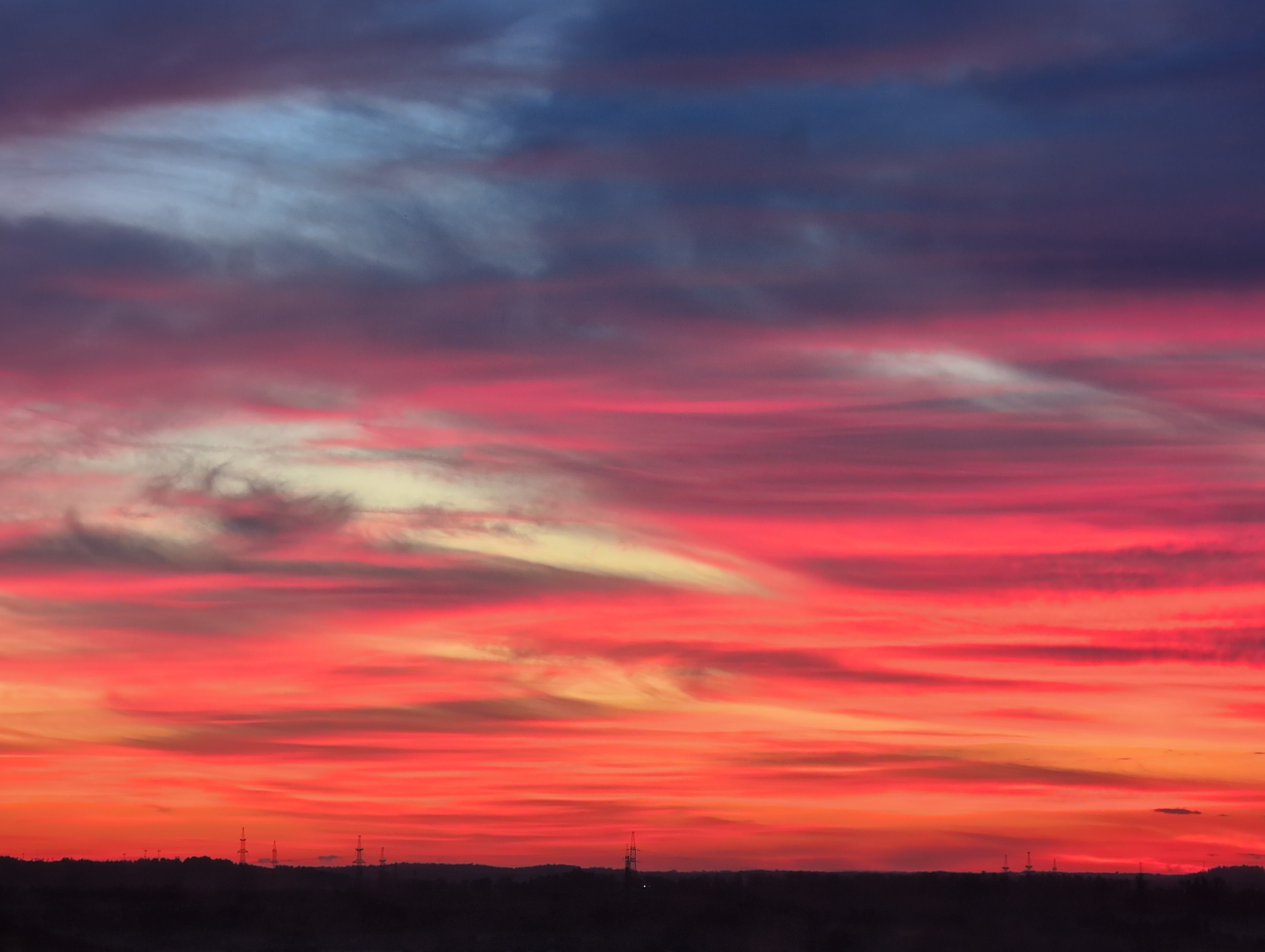 The image depicts a vibrant sunset over the city of Bugry, Russia. The sky is a spectacle of colors, with streaks of deep blue and purple at the top fading into bright pinks and fiery oranges towards the horizon. Wispy clouds are scattered throughout, catching the light and adding texture to the dramatic sky. Silhouetted against the colorful sky is a dark, undulating landscape of trees and rolling hills, characteristic of the Bugry region. Several tall, industrial structures, likely power line towers or similar infrastructure, are visible standing starkly against the horizon line. The overall impression is one of natural beauty and the industrial presence within the landscape.