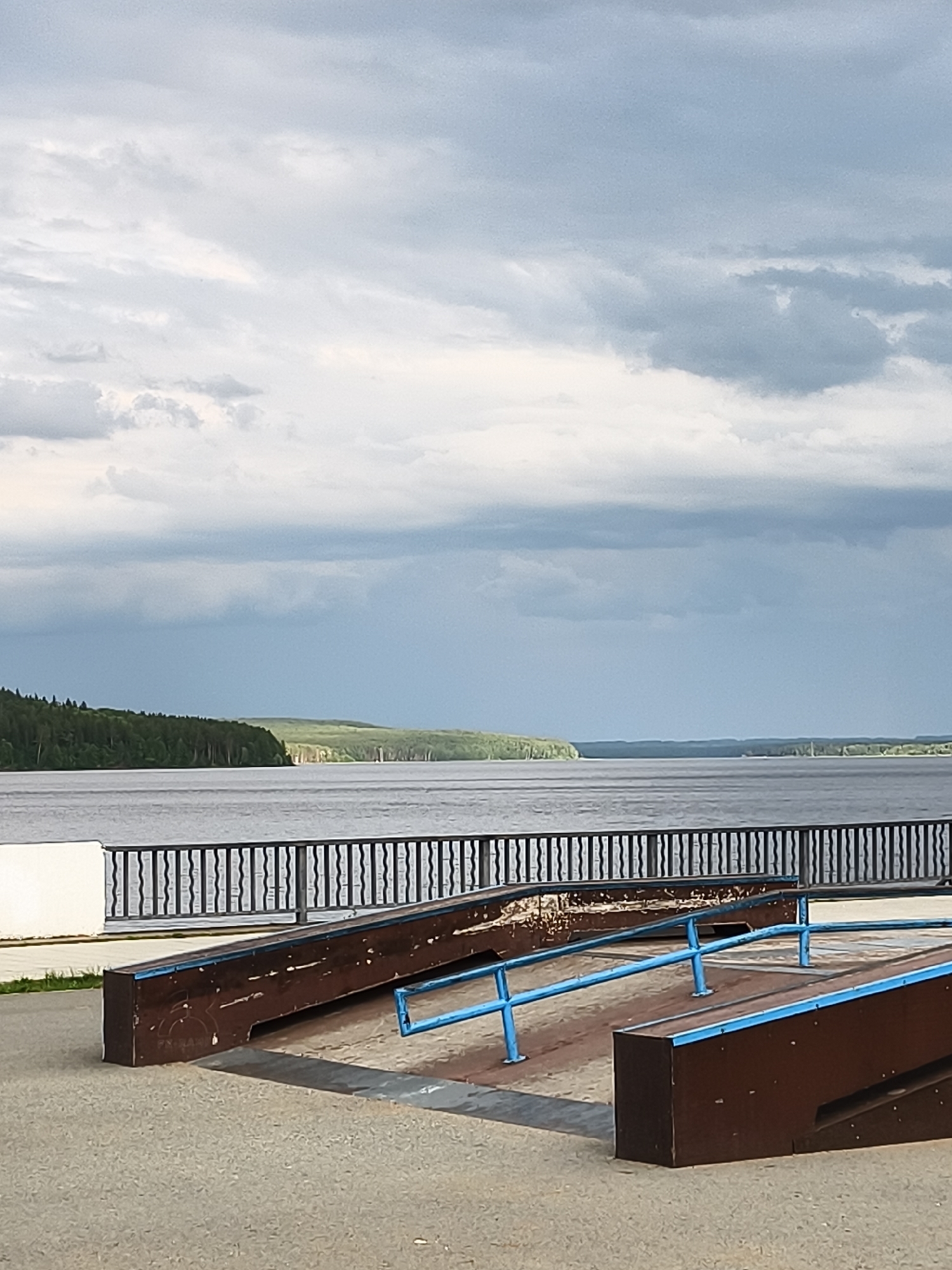 The image captures a view of the Kama River in Chaikovsky, Russia. In the foreground, a skate ramp made of brown wood with blue metal railings is visible. A black metal fence with vertical bars separates the skate area from the riverbank. The river is wide and calm, with a distant shoreline characterized by a dense forest. The sky is overcast with heavy, grey clouds, suggesting a potentially rainy day. The overall scene is one of an outdoor recreational area by a large body of water under a cloudy sky. There are no people or animals present in the image.