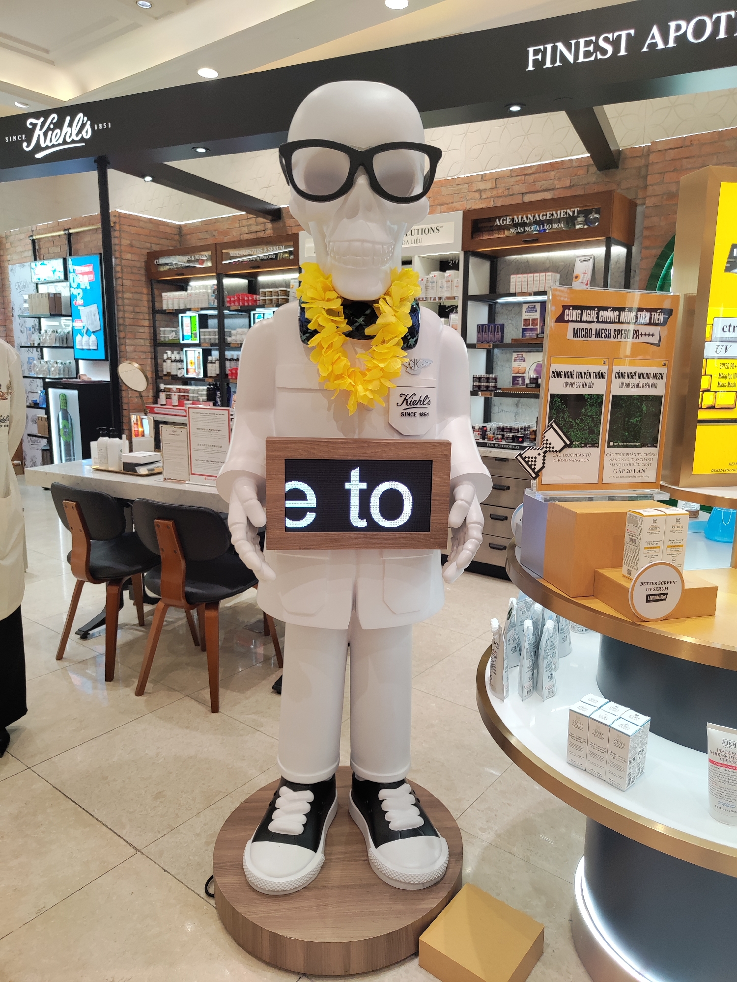 The image captures the interior of a Kiehl's store, likely located within a shopping center or department store. Dominating the foreground is a white mannequin figure resembling a skeleton wearing glasses, a yellow lei, a white lab coat, and black and white sneakers. The mannequin holds a sign that reads "e to". The store's layout includes product displays on shelves and tables, showcasing various Kiehl's skincare and cosmetic products. Behind the mannequin is a checkout counter with chairs. Signs advertising sunscreen and anti-aging products are visible. The store's design includes brick accents and modern lighting. "FINEST APOT" is clearly displayed on the overhead sign. Kiehl's branding is prominent throughout. The context suggests the photo was taken inside a department store in Thủ Đức, Vietnam.