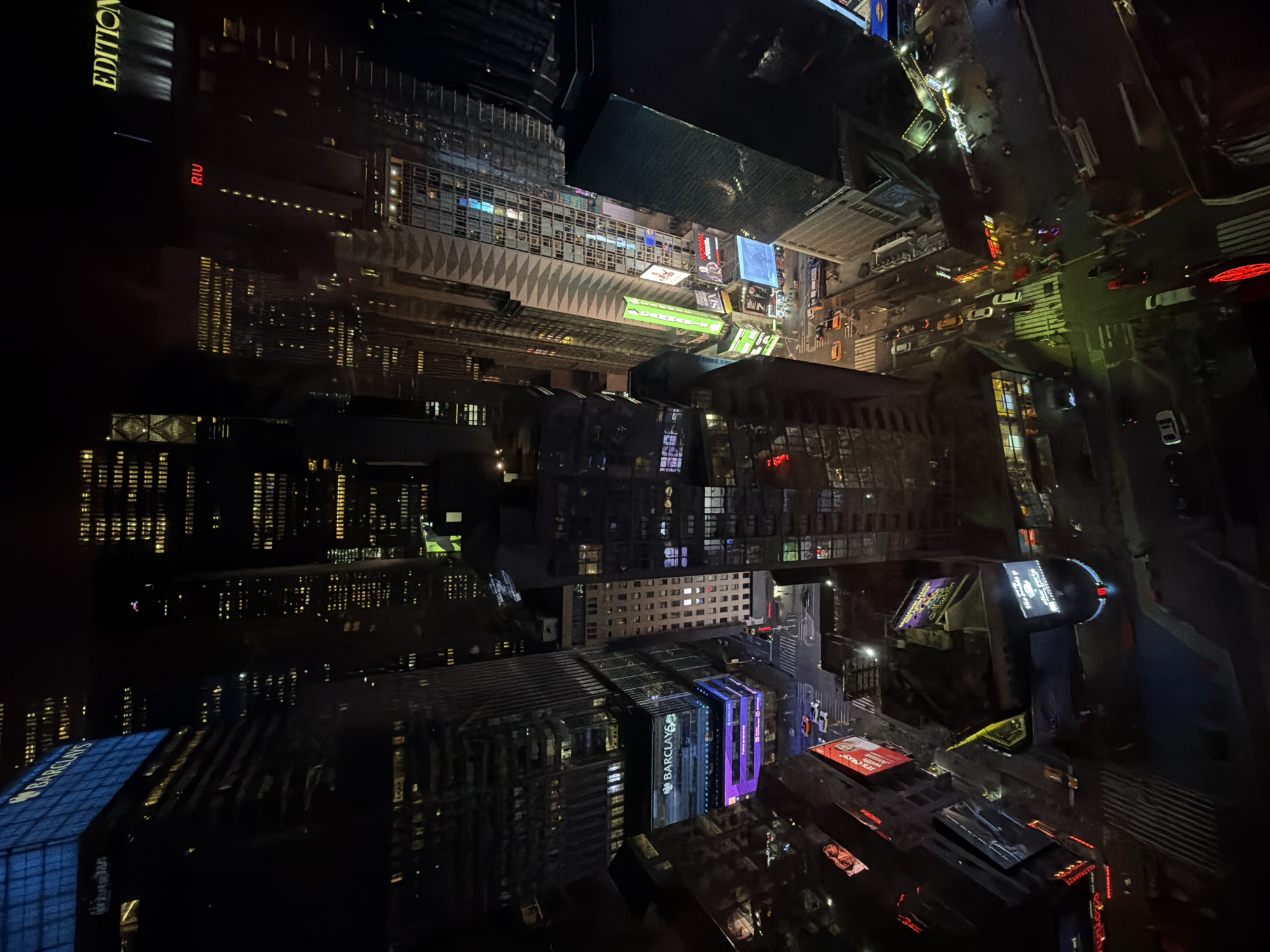 This image captures an aerial view of a city at night, likely Times Square in New York City, United States. The urban environment is filled with towering buildings, many illuminated with various lights and displays. The street below is bustling with vehicular traffic and pedestrian activity, visible through the reflections of light. Signage and advertisements are prominent, indicating a commercial and lively setting. Buildings like the EDITION, RIU, and Barclays are identifiable, adding to the landmark quality of the scene.