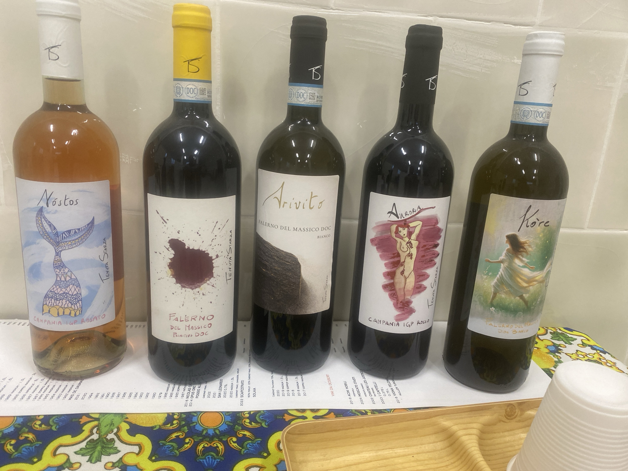 Five bottles of wine are lined up in front of a white tiled wall, likely in the Italian city of Falciano del Massico, as indicated by the regional wine appellations on the labels. From left to right: a bottle of "Nostos" Campania IGP Rosato; "Falerno del Massico DOC Primitivo"; "Arivito" Falerno del Massico DOC Bianco; "Aurora" Campania IGP Rosso; and "Kore" Falerno del Massico DOC Bianco. The bottles are arranged on a surface covered with a printed tablecloth with a floral and citrus fruit pattern. Below the bottles are printed sheets of paper with lists of vintages and wine names. To the right are some empty white cups and a wooden tray.