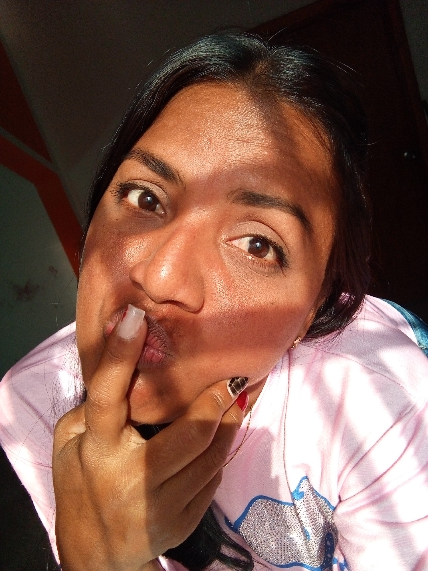 A woman is shown in a close-up selfie. She is wearing a light pink shirt and has dark hair. Her right index finger is touching her lips in a pouting expression. Light is shining on her face, creating shadows. The background shows a glimpse of a door and wall details.