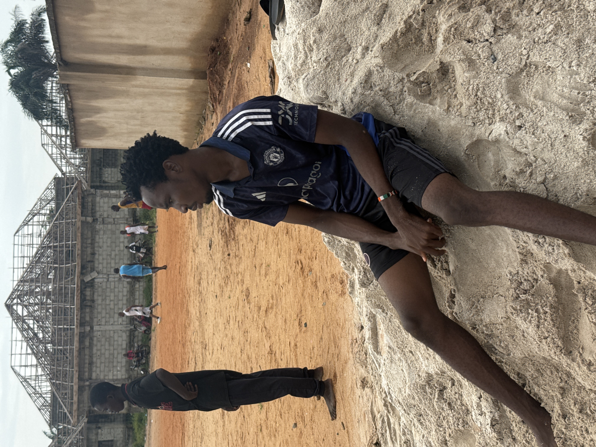 In Ihiagwa, Nigeria, a young Black man with dark, curly hair is seated on a pile of light-colored sand, looking to his right. He wears a dark blue soccer jersey with white stripes on the sleeves and the "Manchester United" crest, along with black shorts. A colorful bracelet adorns his left wrist. His legs are extended, and his hands are resting on his lap. To his left, another young Black person in a black shirt and pants stands barefoot on an orange dirt field. In the background, several other young Black men are visible. Some are walking on the dirt field, and others appear to be near motorcycles. Behind them, a multi-story building with a visible wooden roof structure under construction stands, and a light brown wall borders the right side of the scene, partially obscuring palm trees. The sky is overcast.