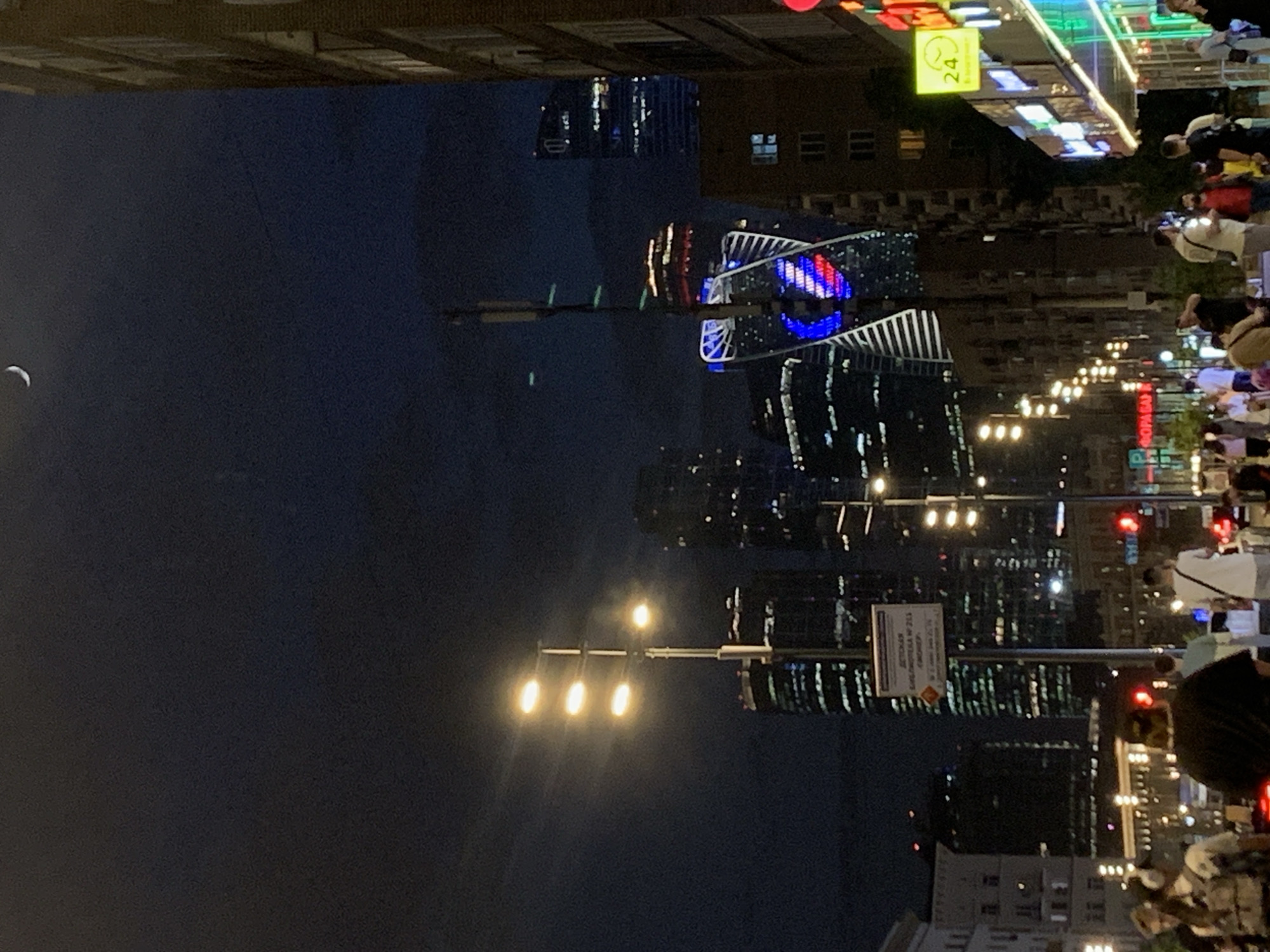 The image depicts an urban night scene, likely taken at dusk or night. The sky occupies the left side of the frame and is dark, with a crescent moon visible in the upper left corner. The right side of the image shows a city street bustling with pedestrian traffic. There are many people walking along the sidewalks. Buildings rise in the background, illuminated with various lights, suggesting a modern urban skyline. Streetlights provide illumination along the road. A sign is visible on a pole, and a barber shop sign with red, white, and blue stripes is also seen. A "24" sign hangs above a building. Traffic signals, including red lights, are visible. The location appears to be a densely populated area.
