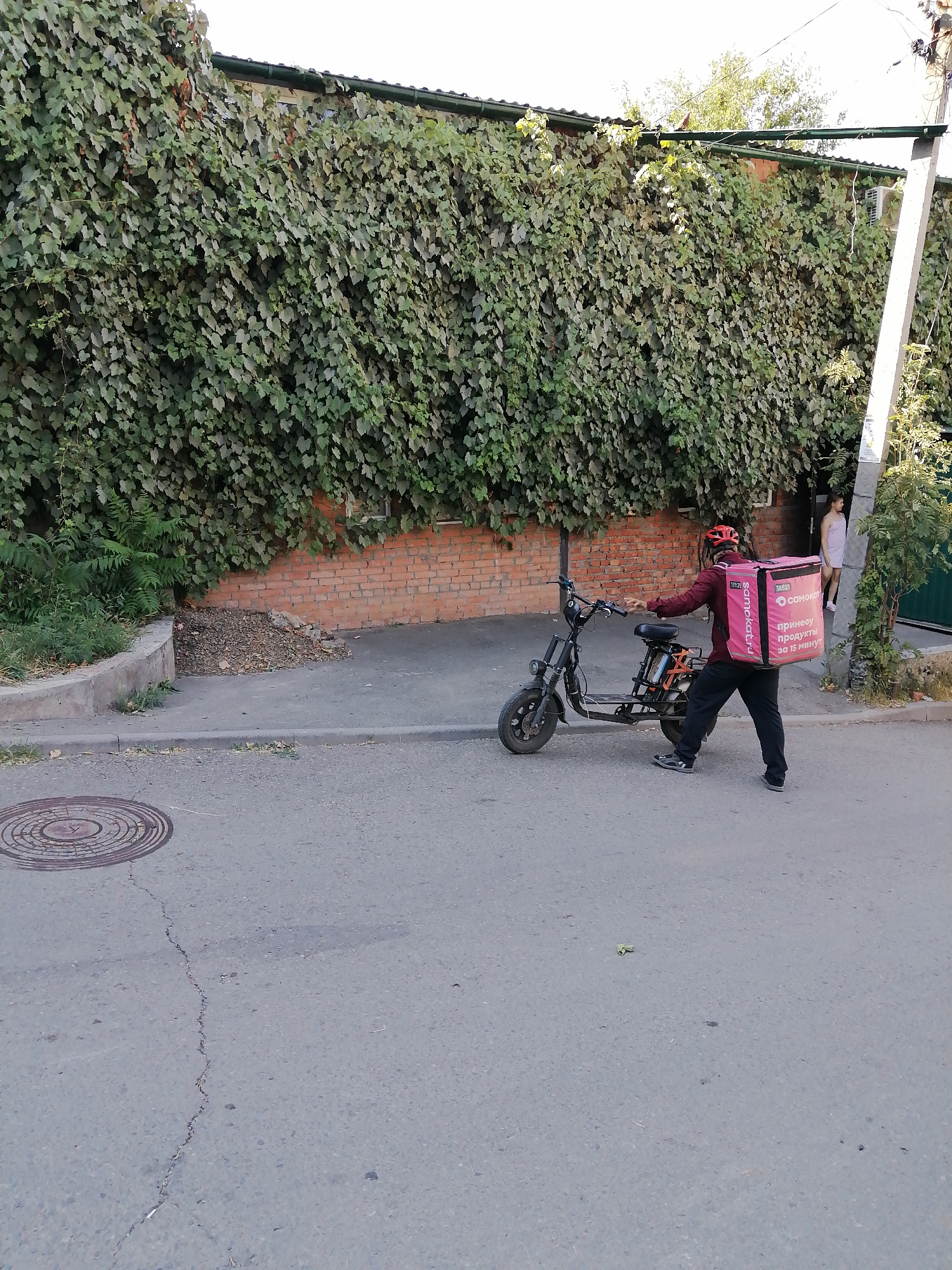 In Rostov-on-Don, Russia, a delivery person wearing a red helmet and a maroon jacket is standing next to a black electric scooter. They are pushing the scooter and have a large pink delivery bag on their back. The bag has the logo "samokat.ru" and Russian text that translates to "bring products in 15 minutes." The background features a brick wall covered in dense green ivy, a concrete sidewalk, and a paved road with a manhole cover. A building with a green roof is visible behind the ivy. To the right, a young woman in a light-colored dress stands near a doorway, and a utility pole with wires is also present. The overall scene suggests a delivery operation in an urban environment.