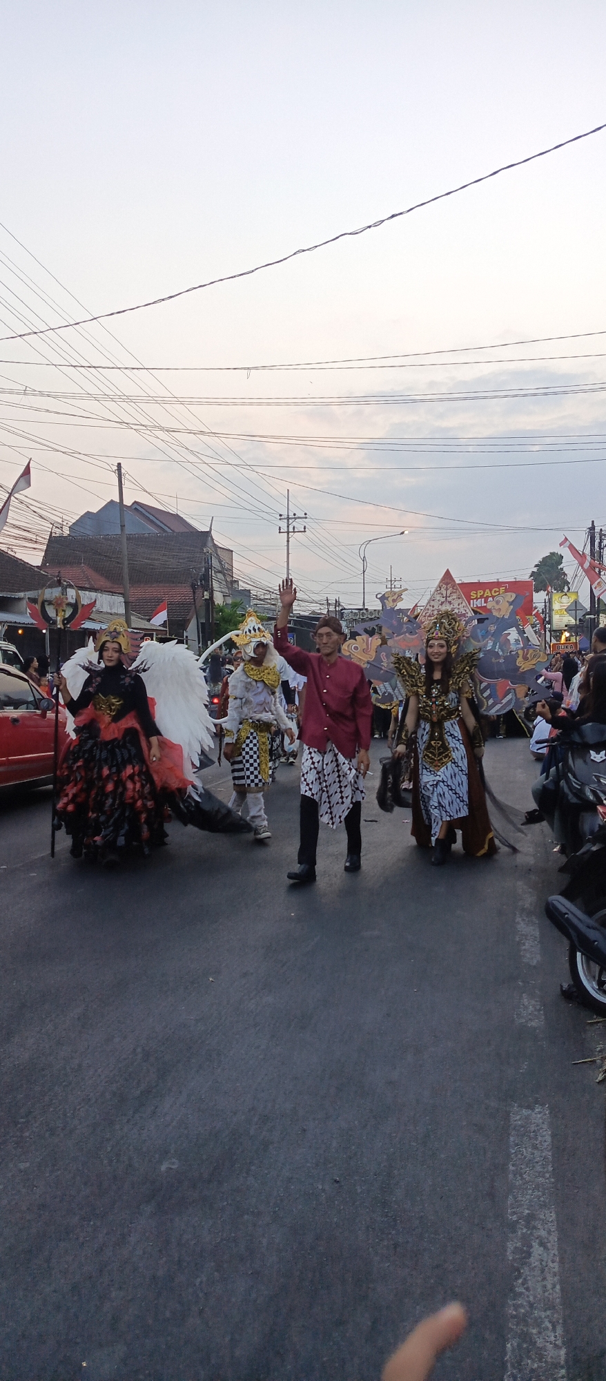 This is a vertical, medium shot of a parade procession on a street in Mangliawan, Indonesia. The sky is overcast with a pale, grayish-blue hue. Several people in elaborate costumes are walking down the street.
On the left, a person in a black and red dress with a large, feathery headdress and white wings is holding a staff. To their right, a person in a white and gold costume with a checkered pattern on their pants is walking. In the center, a man in a maroon shirt and patterned pants is waving to the camera. To his right, a woman in a golden, ornate costume with large wings is smiling. Behind them, a large banner with a red, triangular graphic and text is visible.
The street is asphalt, and there are several parked motorcycles on the right side of the frame. Buildings and power lines are visible in the background. Indonesian flags are also displayed along the street. The overall scene suggests a festive cultural event taking place in the early evening.