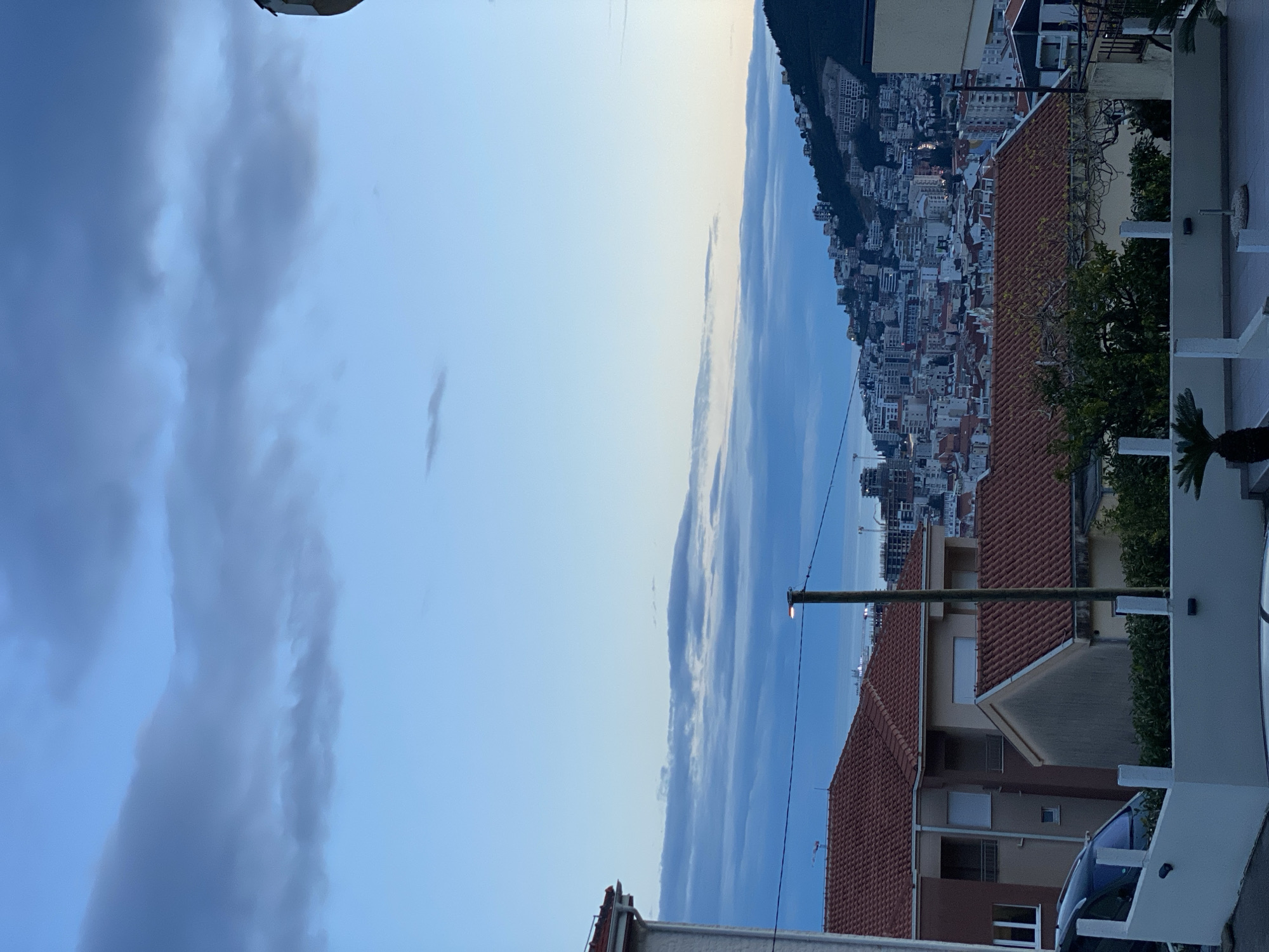 The image is an eye-level, outdoor daytime view of a cityscape, likely Budva, Montenegro, as suggested by the presence of dense urban development near the coastline and a mountainous backdrop. The image is oriented vertically with the sky on the left and the cityscape on the right.
The sky occupies a significant portion of the left side, exhibiting a mix of light blue sky and layered, grey-blue clouds.
On the right, the cityscape consists of closely packed buildings with a variety of architectural styles, likely residential apartments and commercial structures. Red-tiled roofs are prominent among the buildings. The city appears to rise in elevation from the coastline to a wooded hillside in the background. The water between the city and the hill is visible, but is indistinct.
In the foreground, there are parts of buildings and other structures that suggest the vantage point is from a higher elevation within the city. A streetlight is present, along with parts of houses.
There are no people visible in the frame. The overall environment suggests an urban area with a coastal setting.