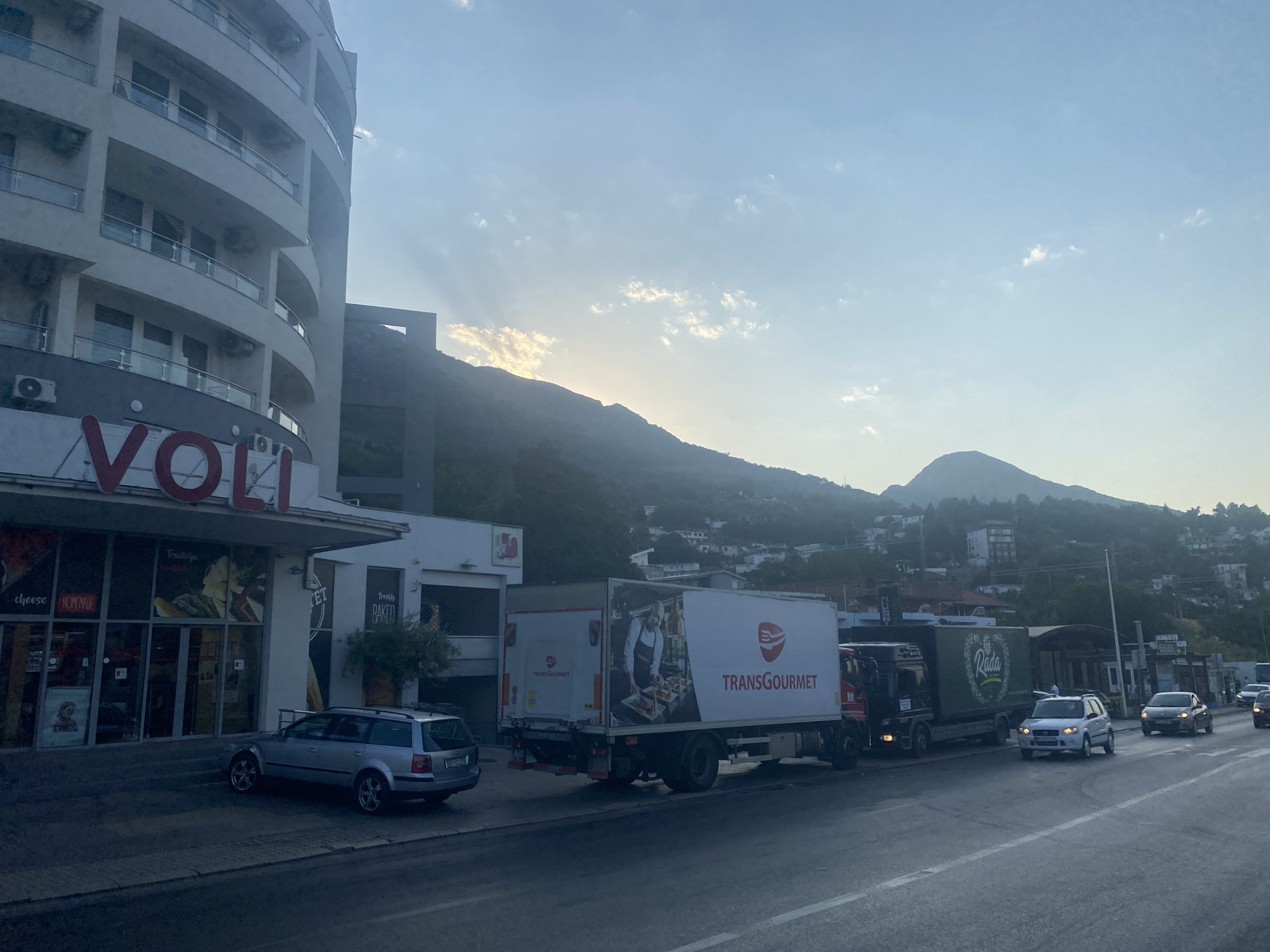 In Sutomore, Montenegro, a scene unfolds by a roadside storefront and its adjacent street. To the left, a multi-story building with a curved facade features balconies with glass railings and multiple air conditioning units. The ground level of this building houses a "VOLI" supermarket, identifiable by large red lettering. The supermarket's large glass windows display various products and promotional signage, including "CHOOSE HOMEMADE" and "FRESHLY BAKED." A silver station wagon is parked in front of the supermarket.
The street in front of the buildings is populated with several vehicles. A large white box truck with "TRANSGOURMET" branding and an image of a person preparing food is parked alongside other trucks. Another truck with a green container bearing the "Rada" logo is also visible. Several cars are on the road, including a white SUV and a darker sedan.
The background reveals a hilly landscape, with numerous houses and buildings scattered across the slopes. The sky above is a pale blue with scattered white clouds, and sunlight filters through the haze, suggesting either dawn or dusk. The overall impression is a view of a commercial area in a coastal town, with daily life and commerce taking place on a street leading towards the mountains.