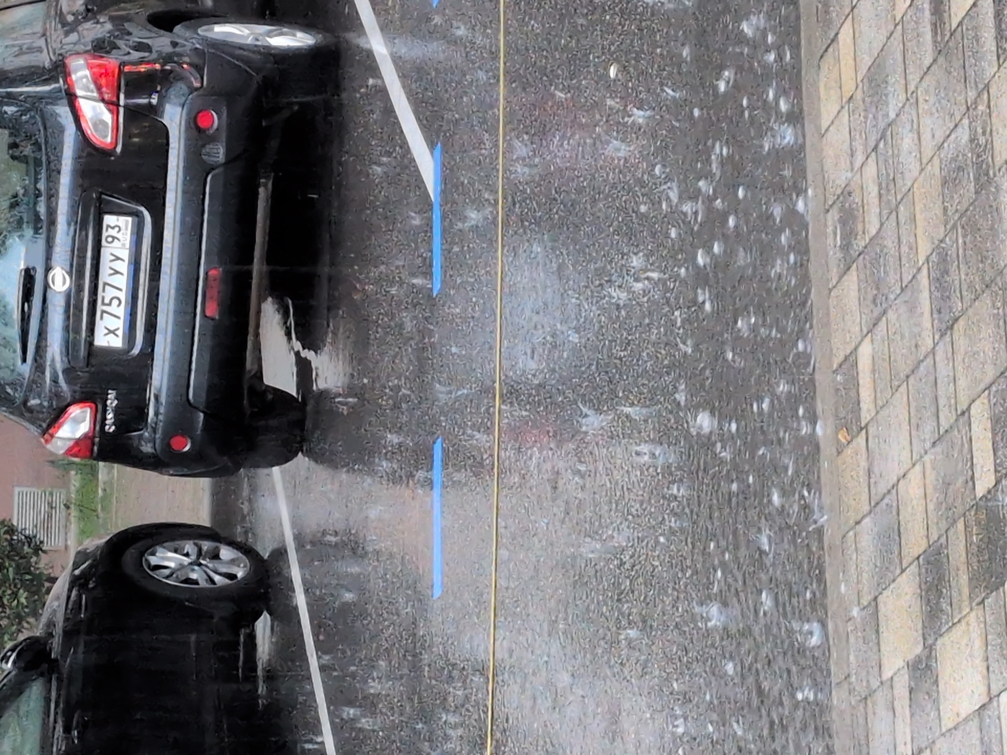 In Sochi, Russia, amidst heavy rainfall, a black Nissan Qashqai is parked on an asphalt surface. The car's rear is visible, with a license plate that reads "X757YY93". The ground is wet and reflects the surroundings. The image also shows the rear of another black car on the left, partially visible. White parking lines are marked on the asphalt, along with two blue rectangular markers. A yellow wire runs horizontally across the lower portion of the image. The scene is a typical streetscape during inclement weather.