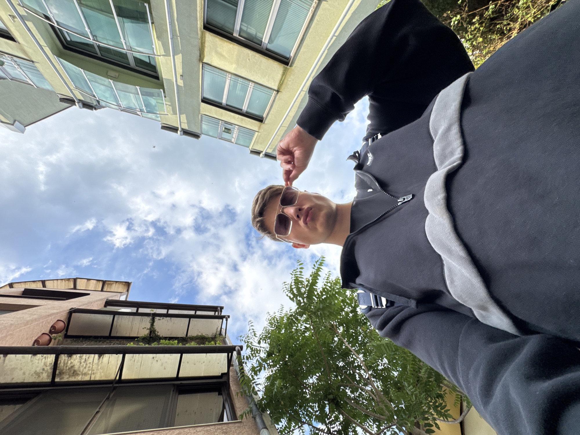 A man wearing sunglasses and a black hoodie with a grey stripe stands beneath buildings and a blue sky with clouds. He is touching his sunglasses. One building is a light yellow color, and the other is brown. Green trees are also visible. The buildings and sky suggest an urban setting. Burgas, Bulgaria is not definitively identifiable from this image.