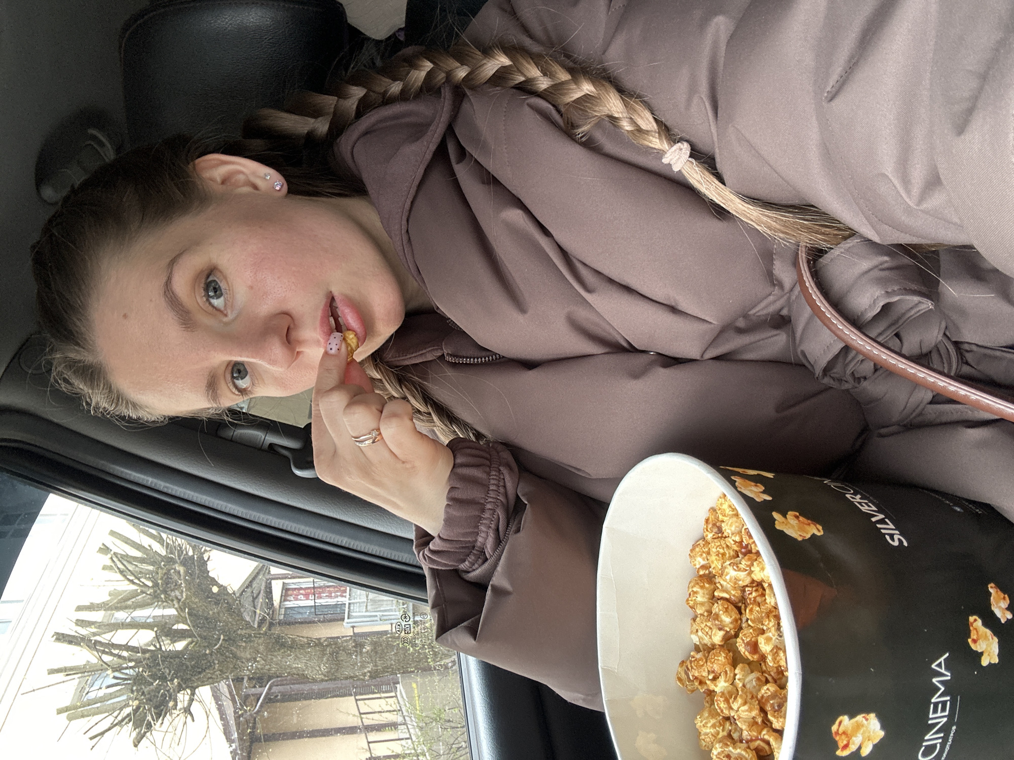 A young woman with fair skin, light eyes, and a blonde braid is eating caramel popcorn in a car. She's wearing a brown puffy jacket. A large black bucket labeled "CINEMA" and "SILVERCINEMA" is partially visible, filled with caramel popcorn. Outside the car window, a bare tree and buildings can be seen. Based on the Silver Cinema brand, the location is Klin, Russia.