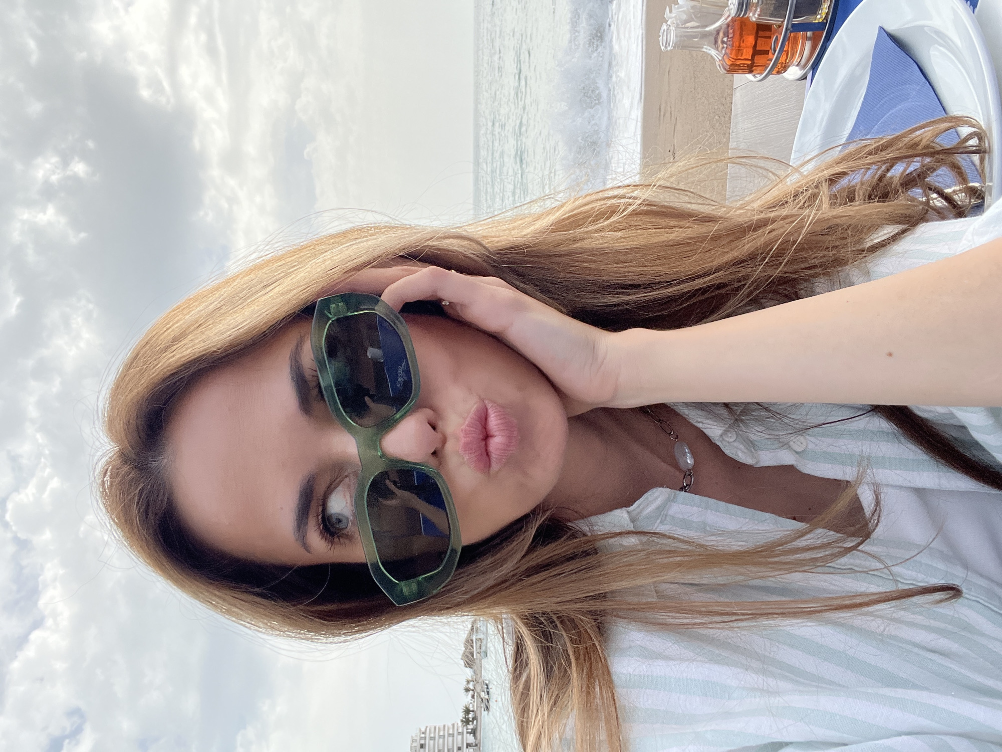 A young woman with long blonde hair and green sunglasses is posing for a selfie. She is pouting her lips and touching her face with her right hand. She is wearing a white and green striped shirt and a necklace with a large pearl. The background shows a beach, the sea, and the sky with clouds. A table with a bottle and a plate is visible in the upper right corner. In the lower left corner, a building and palm trees suggest the location is in a coastal city. Based on the architecture, the location is likely Rafailovići, Montenegro.