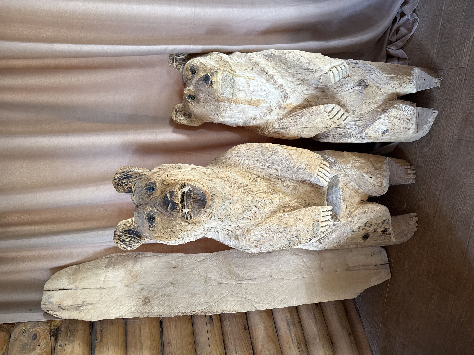 Two carved wooden bear figures lie horizontally on a surface. The bottom bear rests on a wooden plank, which is on a wooden floor. The top bear rests on a light tan cloth. The scene is located in Gornje Mrke, Montenegro.