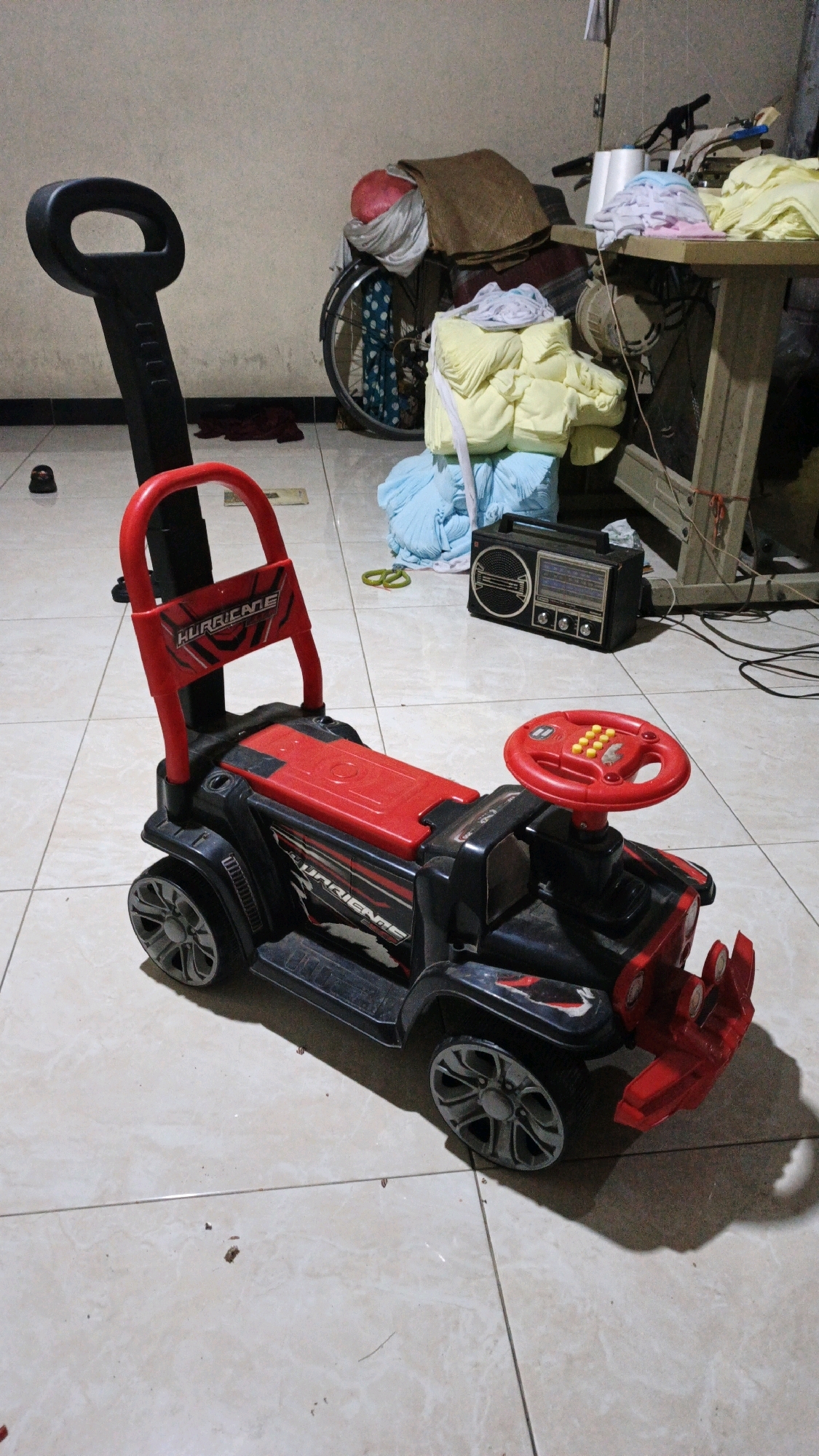 In Waung, Indonesia, a child's red and black ride-on car with a push handle sits on a tiled floor. The car has a red steering wheel with yellow buttons and a "HURRICANE" decal on its side. In the background, a bicycle is partially visible, laden with various items, including fabric and a red ball. A sewing machine with spools of thread and a radio are also present in the cluttered environment.