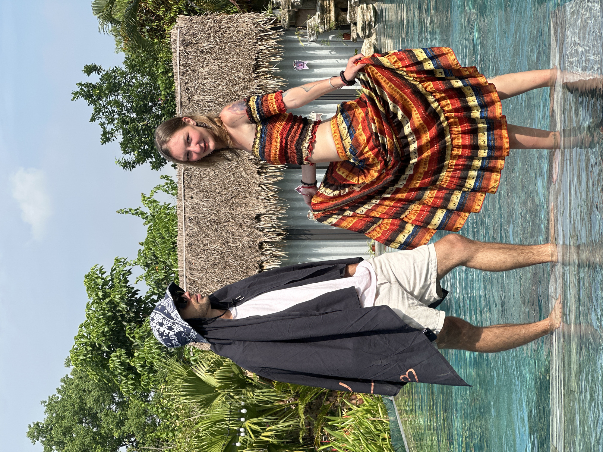 In Nam Hoa Lư Ward, Vietnam, a man and a woman stand in a swimming pool, their lower legs submerged in the clear blue water. The man, on the left, wears a black bandana, sunglasses, a white t-shirt, and light-colored shorts. He is partially covered by a black draped towel. The woman, on the right, sports a vibrant off-the-shoulder cropped top and a matching flowing skirt, both adorned with a colorful striped pattern featuring geometric motifs. Both individuals are facing the camera. Behind them, a thatched-roof structure, possibly a cabana or hut, is visible, surrounded by lush green foliage and trees. The sky above is a clear blue with scattered white clouds. The scene appears to be a relaxed, outdoor setting, likely a resort or a private residence with a pool.