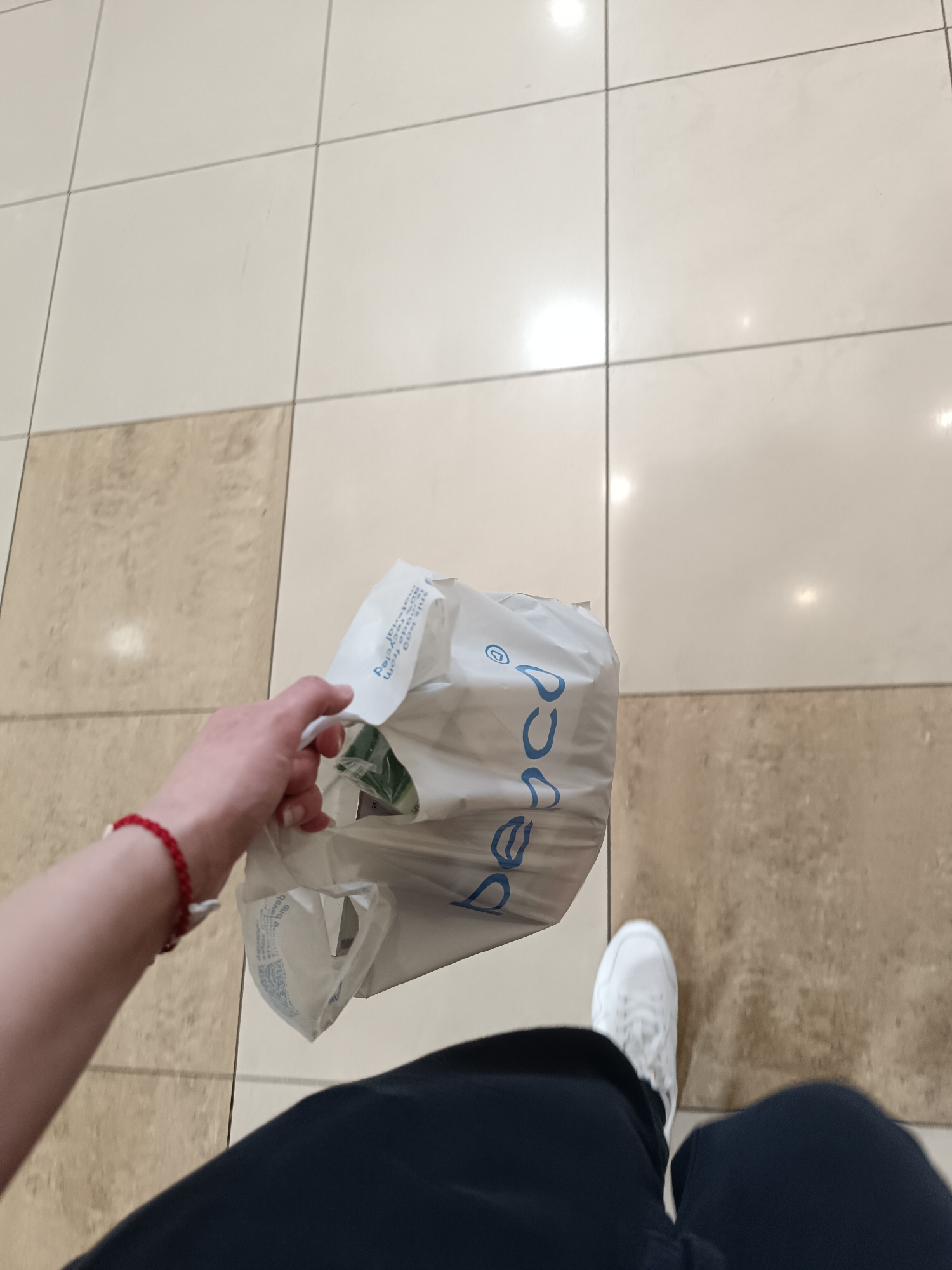 A first-person perspective shot shows a person walking on a tiled floor, holding a white plastic shopping bag from the Bulgarian clothing retailer, "Pepco". The person is wearing dark-colored clothing and white sneakers. A red beaded bracelet is visible on their left wrist. The tiled floor consists of light beige and white squares. The image appears to have been taken inside a shopping mall in Sofia, Bulgaria, given Pepco's presence there.