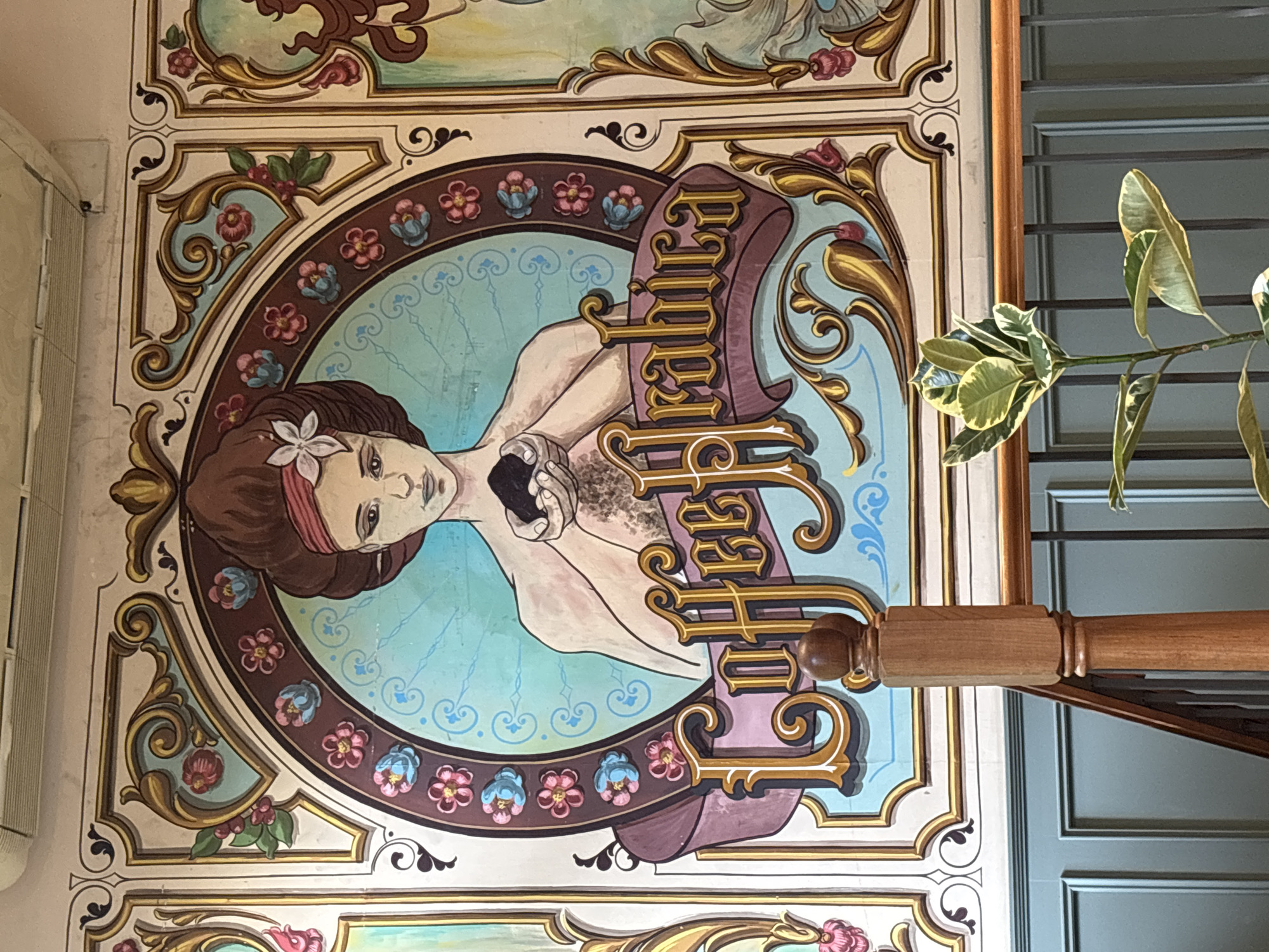 An ornate, vibrant mural adorns a wall or ceiling, featuring a central portrait of a woman. She is depicted from the chest up, with fair skin, brown hair, and a red headscarf or bandana, accented by a white flower near her temple. Her expression is serene and contemplative as she looks slightly upwards. She holds a dark, textured object, possibly coffee beans, against her chest.
The woman is framed within a circular motif, surrounded by elaborate decorative borders that include stylized floral patterns in pink and blue, intricate gold scrollwork, and contrasting light blue and brown backgrounds. The overall artistic style is reminiscent of vintage advertising or traditional decorative art.
Prominently featured within the painting is the text "EL CULTURAL," written in an ornate, golden serif font on a purple-brown banner. The final letter "L" of "CULTURAL" is partially obscured by a wooden pole that extends horizontally across the image. This pole, possibly part of a railing or banister, is made of polished light brown wood with carved details.
To the right of the wooden pole, a plant with variegated green and white leaves is visible. Behind the pole and plant, a section of a light blue wall with horizontal paneling or blinds can be seen. The image is taken from a low angle, looking upwards at the artwork, which is likely found in an interior setting in Buenos Aires, Argentina.