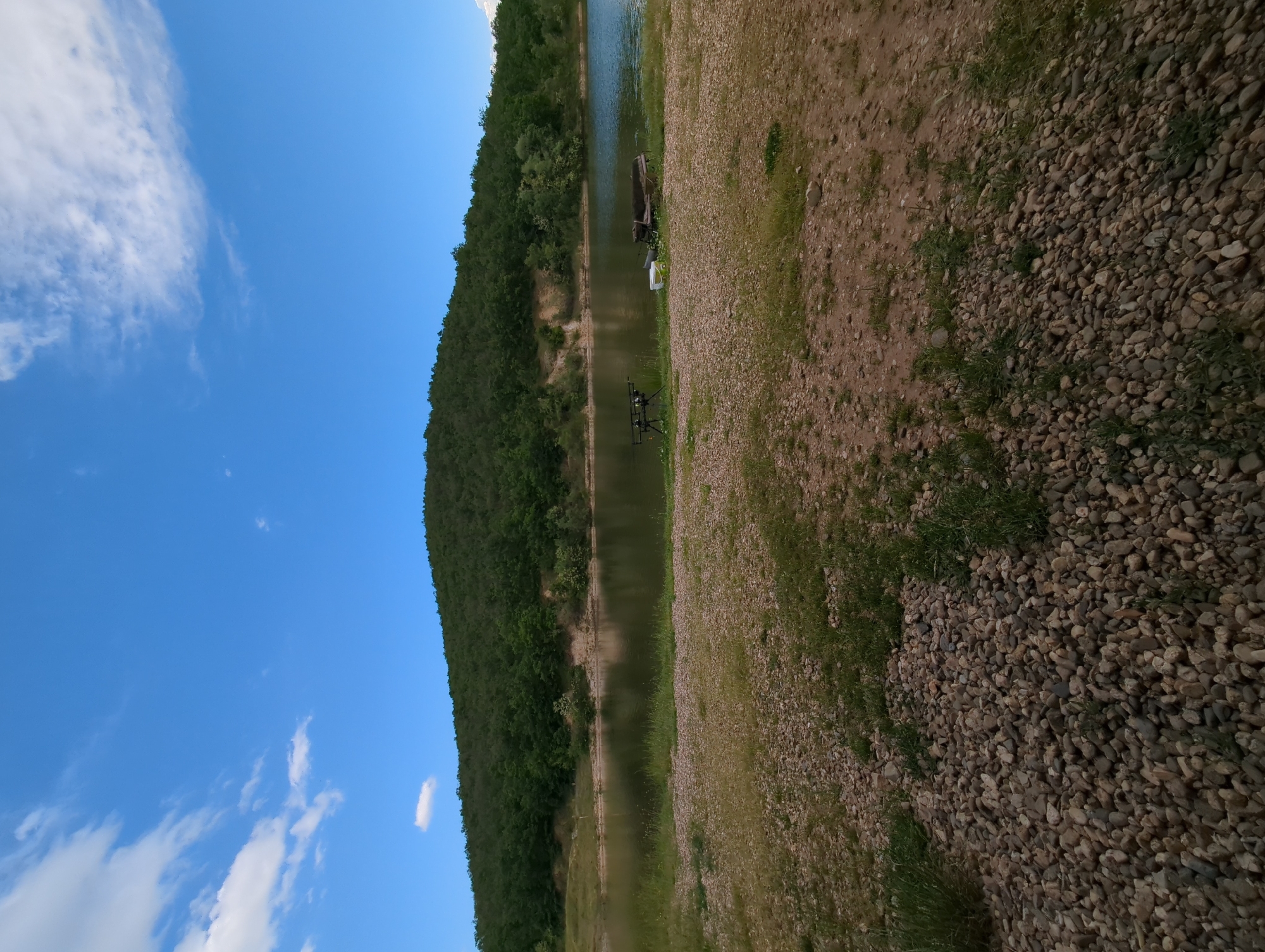 The image depicts a landscape near Slavovitsa, Bulgaria, featuring a body of water bordered by a densely wooded hill on one side and a gravelly embankment with sparse vegetation on the other. In the water, a small boat and a fishing rod stand are visible. The sky above is blue with scattered clouds.