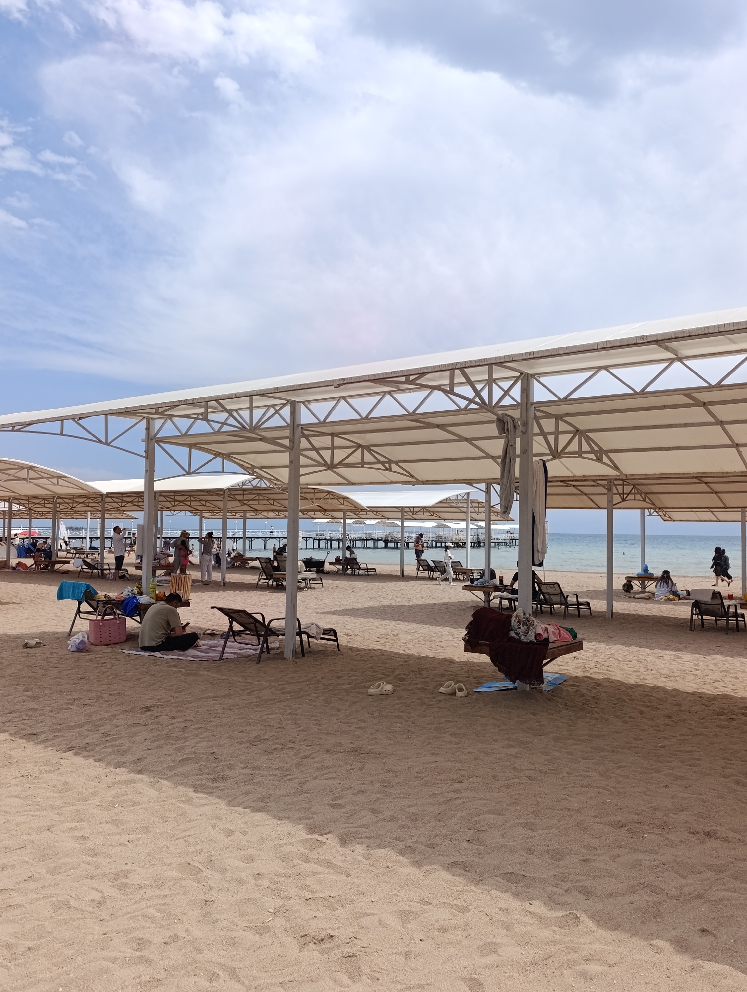 The image depicts a beach scene in Sary-Oy, Kyrgyzstan, featuring a sandy expanse with numerous white canopies providing shade. Several lounge chairs are positioned on the sand, some occupied by people. In the foreground, a person sits on a blanket on the sand, looking at their phone, with a pink bag and a blue towel nearby. Further back, other individuals are visible standing or sitting under the canopies, engaged in various activities. A wooden pier extends into the calm, blue water of the lake, with more canopies visible in the distance. The sky is partly cloudy, with white and gray clouds against a light blue backdrop. The overall impression is one of a relaxed, daytime beach environment.