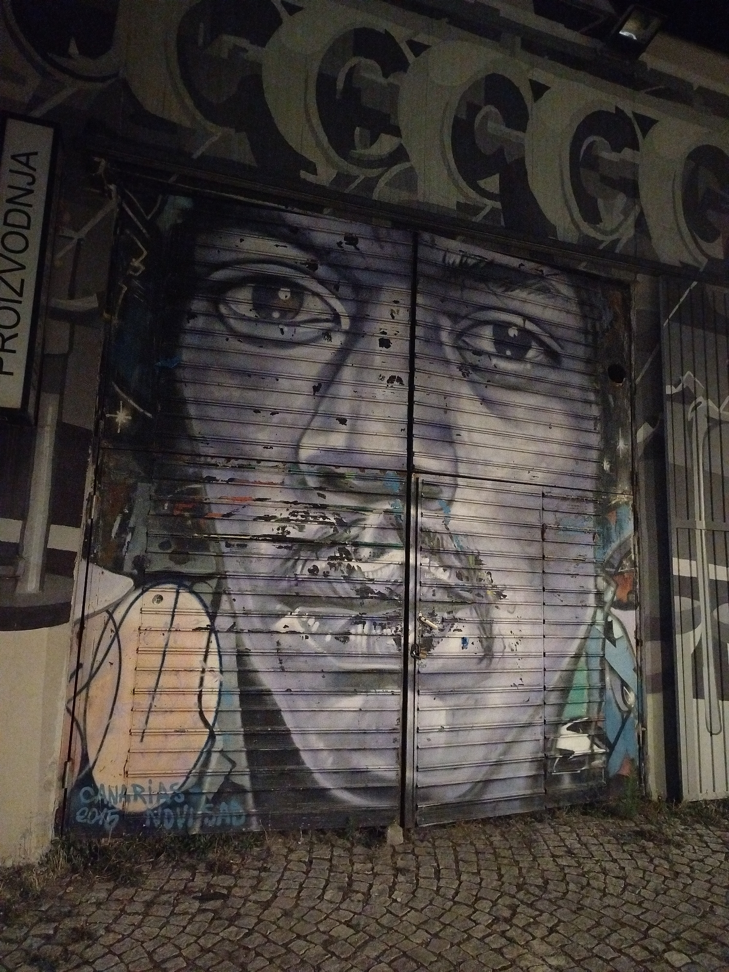 A mural of a face with closed eyes and a mustache is spray-painted on a corrugated metal rolling door in Novi Sad, Serbia. The face is rendered in shades of gray and blue, with some areas of color appearing to be peeling or damaged. To the left of the rolling door is a sign that reads "PROIZVODNJA," which means "production" in Serbian. Below the mural, graffiti reads "CANARIAS E.A.D." and "2015 NOVI SAD." The rolling door is part of a building, and the ground in front of it is paved with cobblestones. The overall scene suggests an urban environment with street art.
