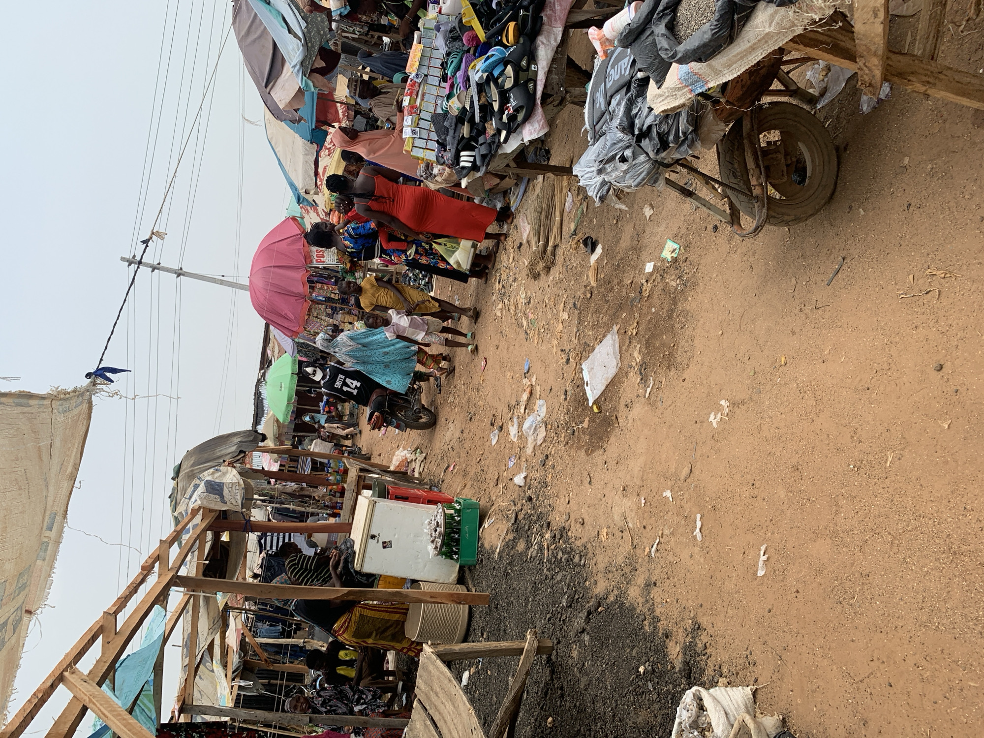 This image captures a vibrant, busy outdoor market scene in what appears to be Keffi, Nigeria, given the contextual information. The scene is set on a dusty, unpaved ground that serves as a thoroughfare, lined on both sides by makeshift market stalls constructed from wooden frames and covered with various fabrics and tarps for shade.
Numerous people are visible, including shoppers and vendors, moving between the stalls or attending to their goods. Many are dressed in casual attire; one woman in the foreground wears a bright red dress, and others are seen with traditional patterns. Children are also present.
The stalls display a variety of merchandise, including shoes (sandals and slippers), clothing, bundled brooms, and sacks filled with various goods. Overhead, power lines crisscross the sky, supported by a utility pole. A large pink umbrella and a smaller green one provide additional shade to vendors or customers.
Towards the right, a rugged wheelbarrow or handcart is loaded with sacks, possibly for transporting goods. Further back on the right, a white cooler/refrigerator and green crates filled with bottles suggest the sale of beverages. The ground throughout the market area is littered with plastic bags, papers, and other debris, indicative of a bustling commercial environment.
The sky is overcast, suggesting a cloudy day, though the time appears to be daytime. Visible text includes "POS" on a sign, "GUP" on a sack, "SMITH 14" on a jersey worn by an individual, "JANG" on a bag, and "CHIPEN" on another sack. The overall impression is one of a lively, everyday market, integral to the local community.