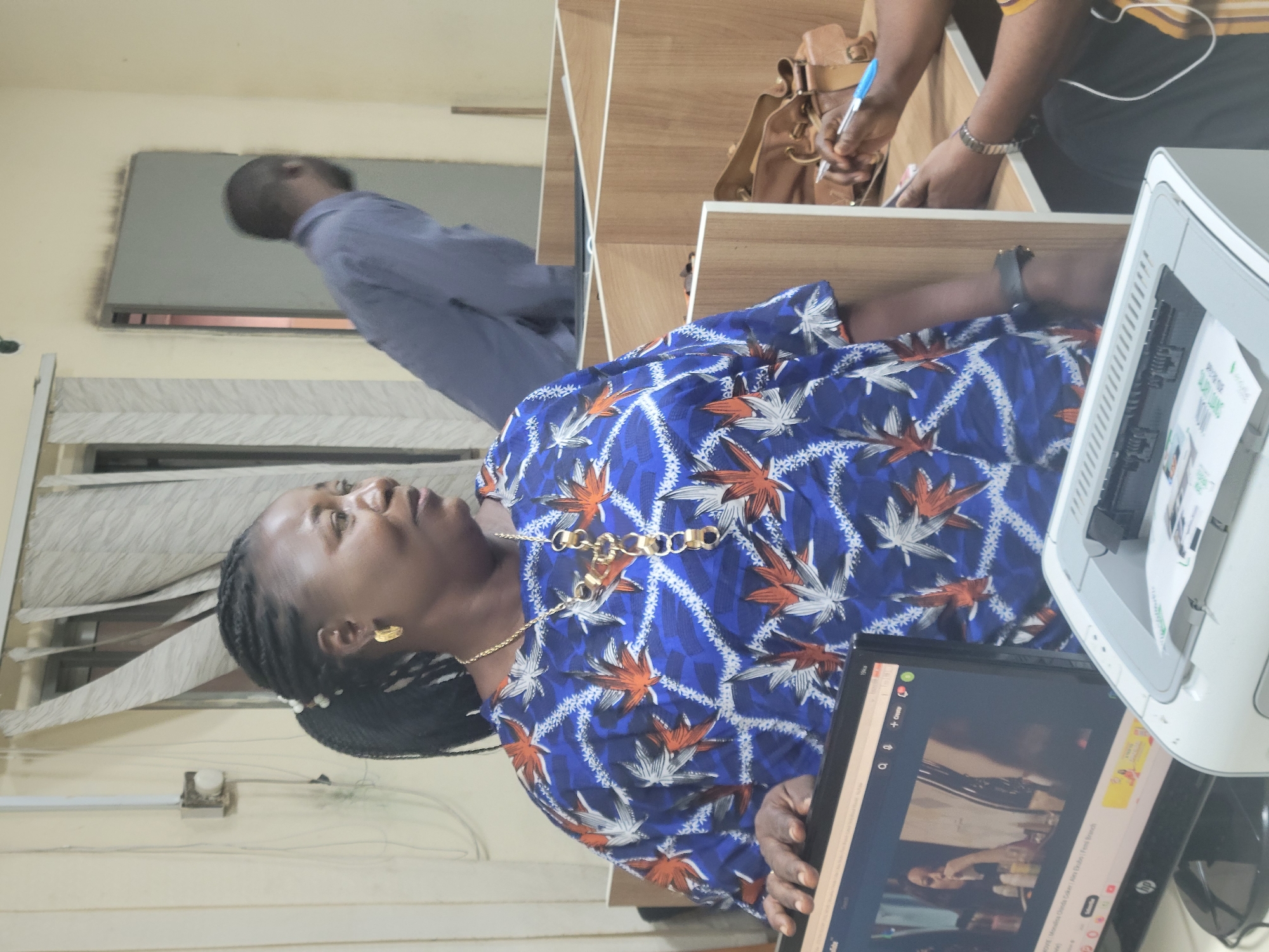 A woman is working at a desk in an indoor office setting in Abuja, Nigeria. She is wearing a blue and white leaf-patterned shirt and a gold necklace and earrings. She is looking up and to the left. In front of her is a laptop displaying a video. To the right of the laptop is a white printer. In the background, another person is writing at a wooden desk. A third person is standing in the background near a window.