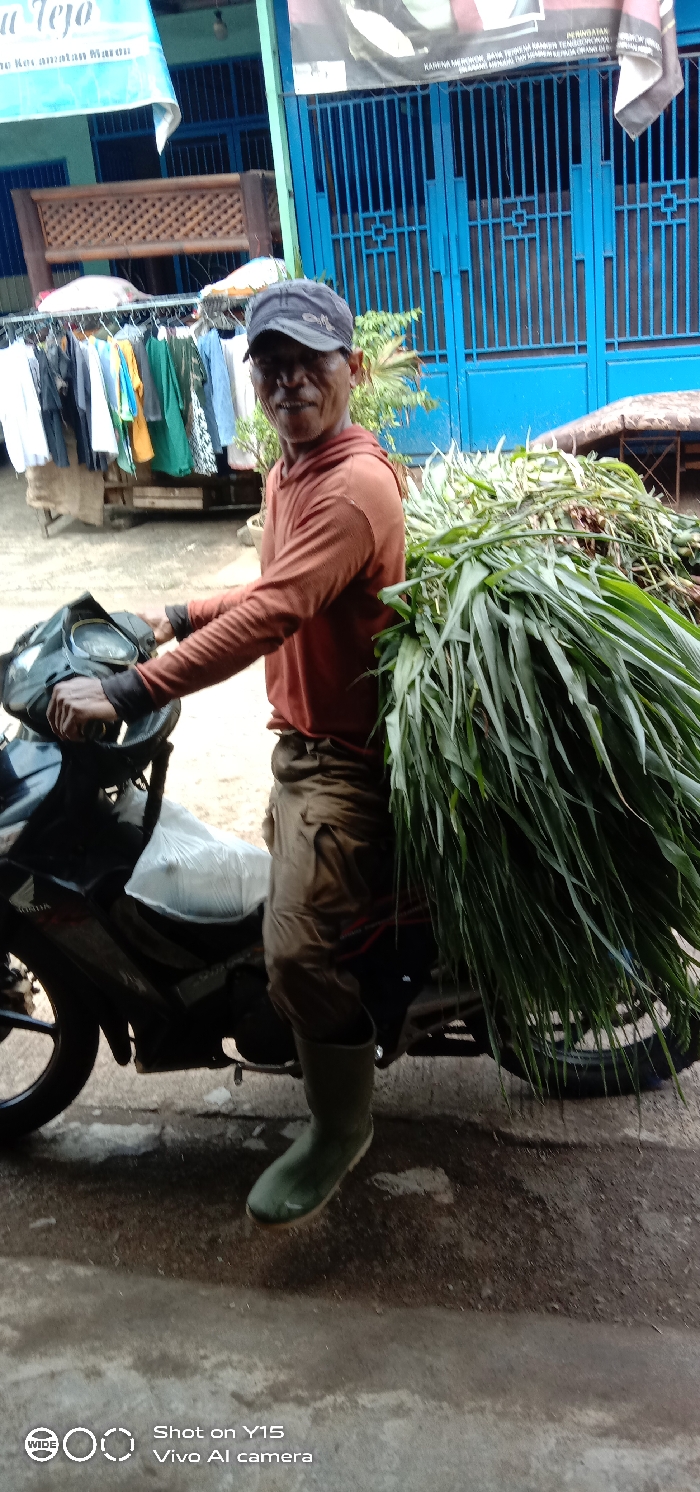 A man on a motorcycle is transporting a large bundle of green grass. He wears a brown long-sleeved shirt, khaki pants, and green rubber boots. A dark cap covers his head. The motorcycle is dark in color. The background includes a blue building with barred windows, and clothes hanging on a line. The ground appears wet and the photograph is taken in daylight.