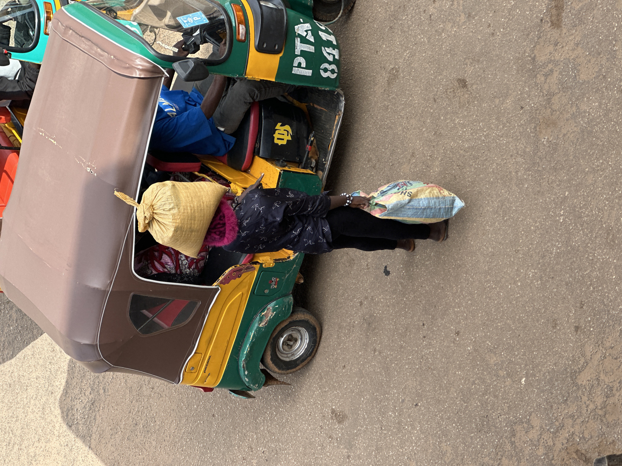 The image depicts a street scene in Bukuru, Nigeria, featuring a yellow and green auto-rickshaw with a brown canopy. A person, wearing dark clothing with a pink fuzzy collar, stands beside the auto-rickshaw, carrying a large, light-colored burlap sack on their head. Another person, dressed in a blue shirt, is seated in the driver's position of the auto-rickshaw. The auto-rickshaw has the registration "PTA 841". The ground is paved asphalt. The person standing is also holding a multi-colored bag. The scene suggests a moment of transit or a stop along a journey within the city.