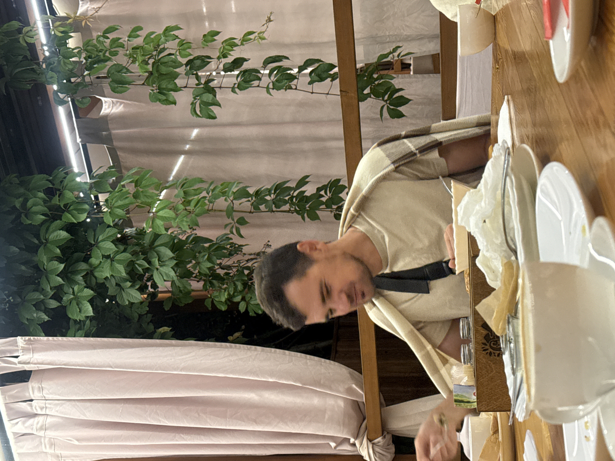 A man with short, dark hair and a small goatee is seated at a wooden table, looking down to his left. He is wearing a light-colored t-shirt and has a plaid blanket draped over his shoulders. A black strap is visible across his chest. In front of him, on the table, are various items including a white cup and saucer, plates with remnants of food, crumpled napkins, and a wooden box. The scene appears to be outdoors, possibly on a patio, with a light-colored curtain and lush green vines visible in the background. The lighting suggests it is evening or nighttime. The location is identified as Orlinovsky District, Ukraine.