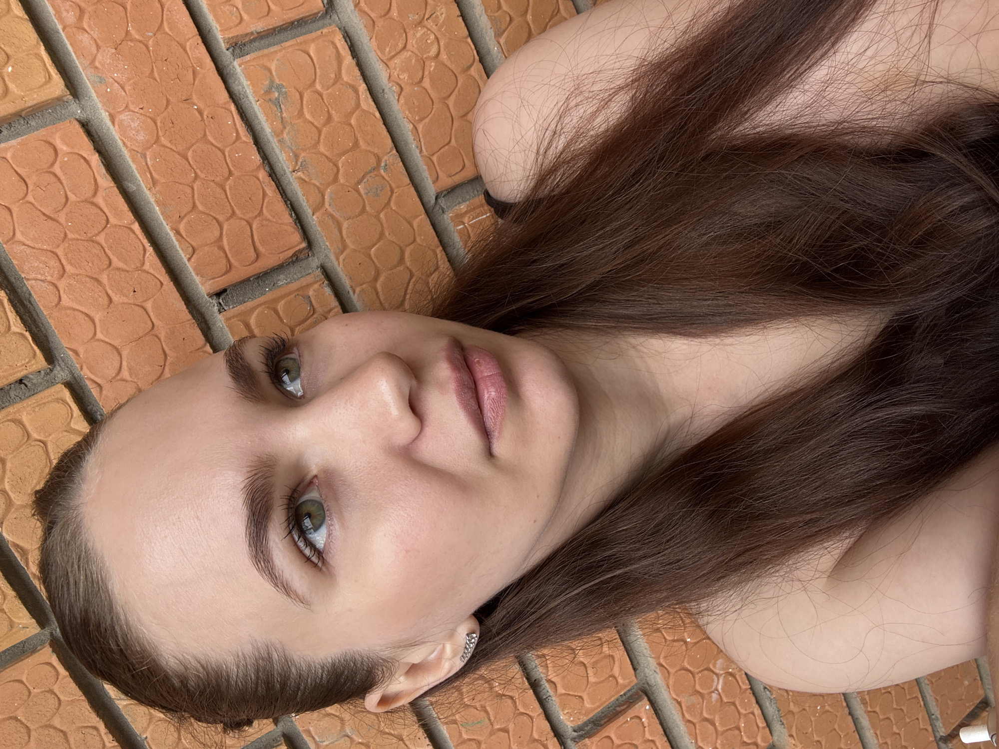 A close-up, high-angle shot captures a young woman lying horizontally on a patterned, reddish-orange brick or tile surface. Her head and upper torso are visible, with her long, dark brown hair fanned out around her. She has fair skin, light green or hazel eyes, and softly colored pink lips. Her expression is neutral and calm, gazing slightly upwards or towards the camera. A small, silver-toned earring adorns her visible left ear. The background is entirely composed of the textured brick pattern, suggesting an outdoor setting, possibly a path or patio. The lighting indicates daylight, with soft, even illumination on her face and the ground. There are no other discernible objects or visible text in the image. The scene is set in Chaikovsky, Russia.