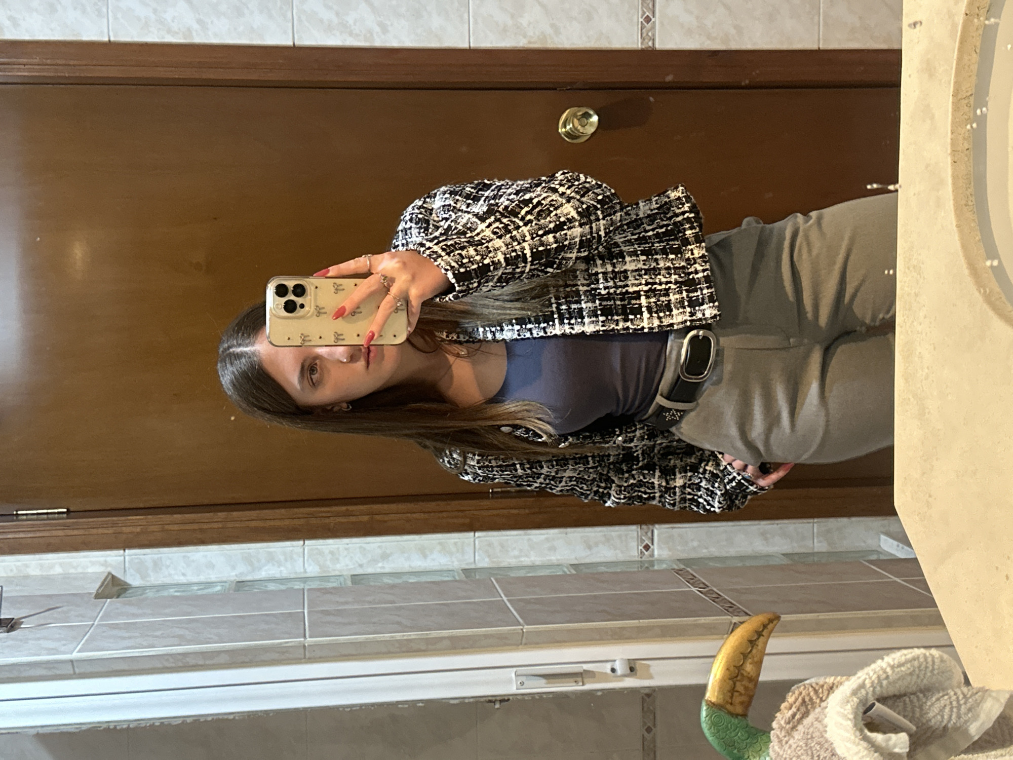 A young woman stands in a bathroom and takes a mirror selfie with her cell phone. She is wearing gray pants, a dark blue top, and a black-and-white tweed jacket. The bathroom features brown tile backsplash and a wood door. The location is implied to be Huixquilucan de Degollado, Mexico.