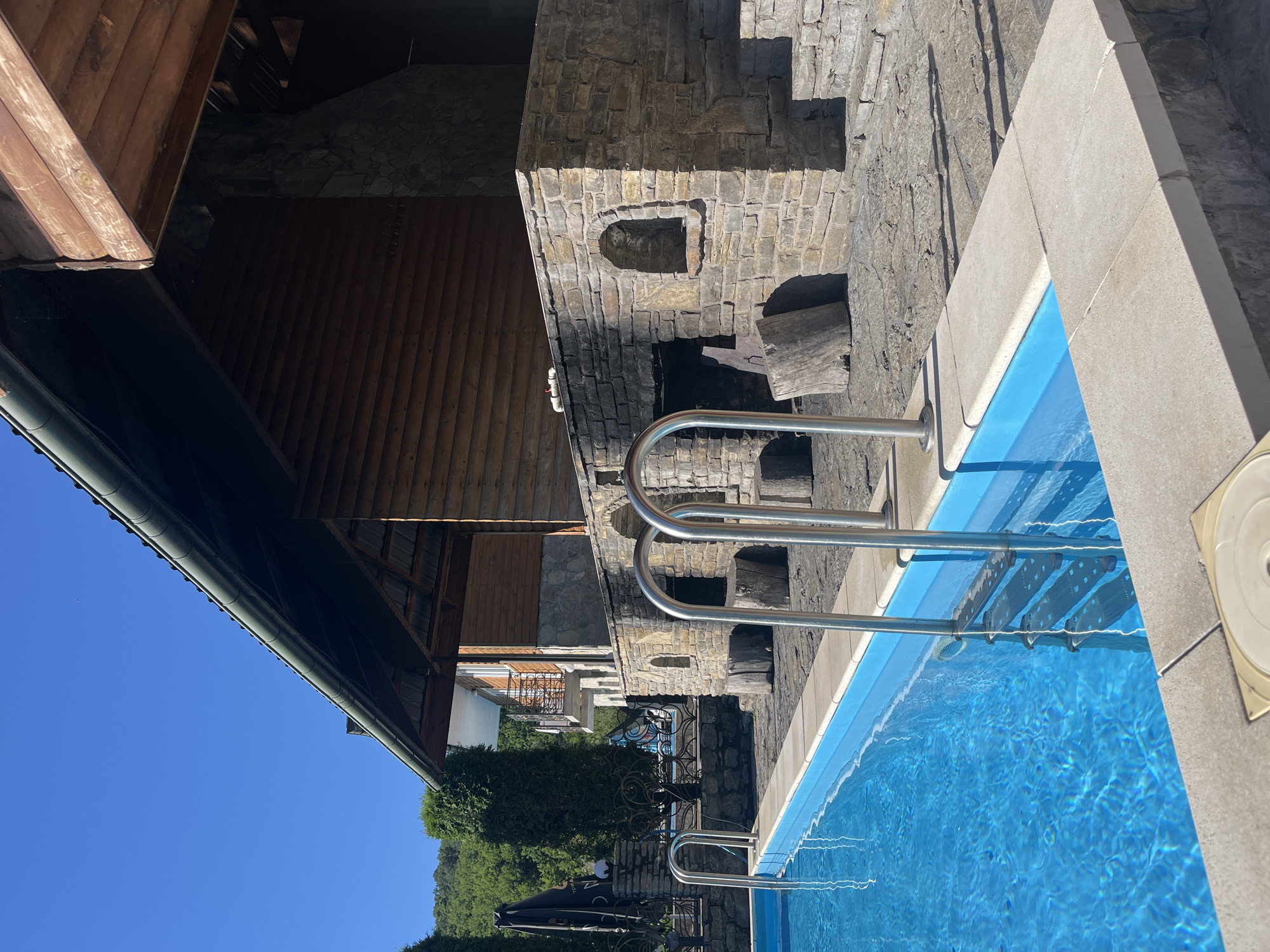 In Vrublivtsi, Ukraine, an outdoor swimming pool area is depicted on a sunny day. The foreground features a bright blue swimming pool with silver metal railings and steps. The pool is surrounded by a concrete deck. Behind the pool, a stone structure resembling an outdoor kitchen or grill area is visible, constructed with rough-hewn stones and featuring arched openings and built-in shelves. Adjacent to this stone structure is a wooden building with a dark roof and log siding. Trees and foliage are present in the background, and the sky is clear blue. There are no people or animals in the frame, and no actions are being performed.