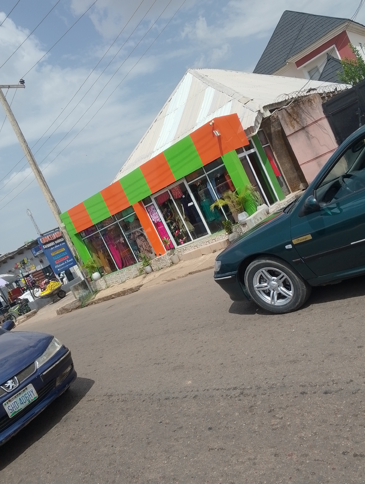 The image shows a street scene in Jos, Nigeria. A green and orange striped building stands prominently in the background, likely a retail store. In front of it, a dark green Peugeot car is parked at an angle. A second blue car, also a Peugeot, is parked on the other side of the road, its license plate reading "SHD-406HY". A sign next to the striped building reads "GRACELAND FASHION WORLD". Near the sign, there's a fruit vendor with a wheelbarrow of yellow fruits, and bicycles are parked nearby. The street appears paved, and overhead utility lines are visible. In the background, a two-story building with a dark grey roof is seen behind the striped building. The sky is bright with scattered clouds.