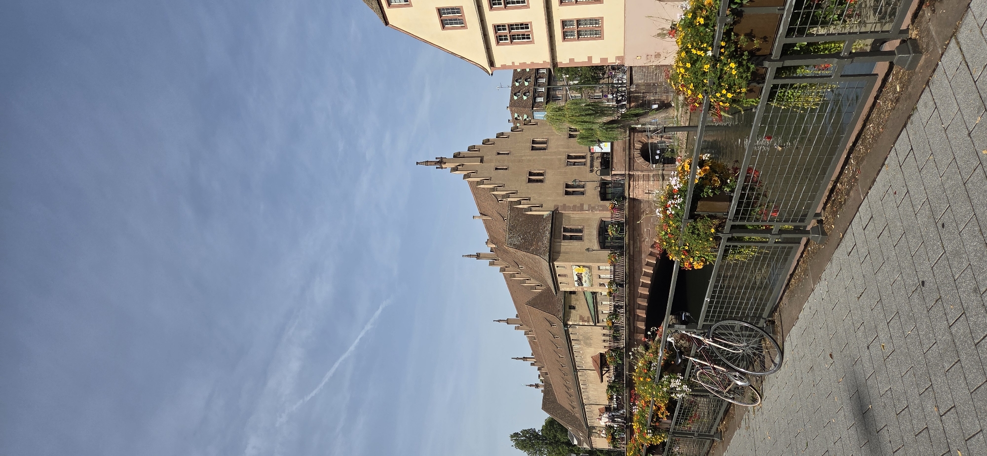 This is a daytime, eye-level, medium shot of the Petite France district in Strasbourg, France. The scene captures a canal with a stone bridge adorned with flower baskets. On the left, a grey paved walkway runs alongside the canal, with two bicycles leaning against a metal railing. The railing is also decorated with hanging flower baskets filled with yellow, red, and white flowers.
In the background, a long, multi-story building with a distinctive steep, gabled roofline and numerous dormer windows stands prominently. The building is made of a light brown material. A smaller, yellow building with red trim and similar window styles is visible to the right of the main building.
The sky is blue with scattered wispy white clouds, one of which forms a contrail. The water in the canal reflects the buildings and the sky. The overall atmosphere is serene and picturesque, typical of a European city's historic canal district.