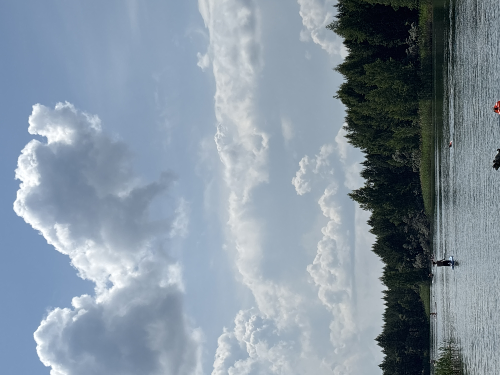 This is an outdoor, full-shot, vertical-orientation, eye-level photograph of a lake in Novyi, Russia on a cloudy day. The sky is filled with large, fluffy white clouds against a pale blue background. The lake surface is rippled, and a dense line of green trees forms the background on the far shore. Several people are visible on the lake: one person is paddleboarding in the foreground, facing away from the camera, while others are further in the distance, also paddleboarding or swimming. Two orange buoys are visible near the bottom of the frame.