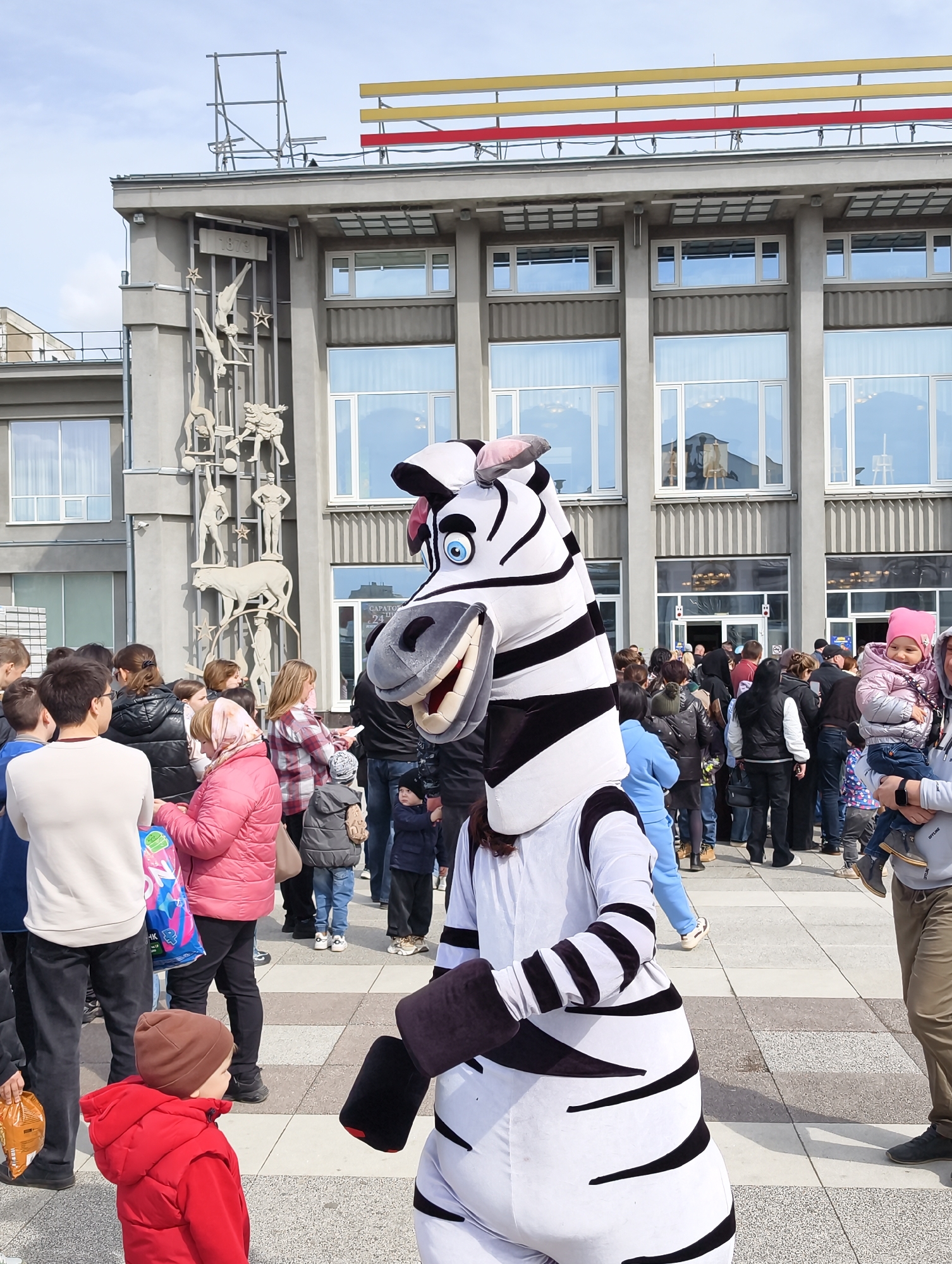 A zebra mascot stands in a public square in front of the Saratov State Circus building in Saratov, Russia. The mascot, wearing a black and white zebra costume, is surrounded by a crowd of people, including children and adults. The building behind the mascot is a large, gray, concrete structure with large windows and a sculpture on its facade. The sky is overcast.
