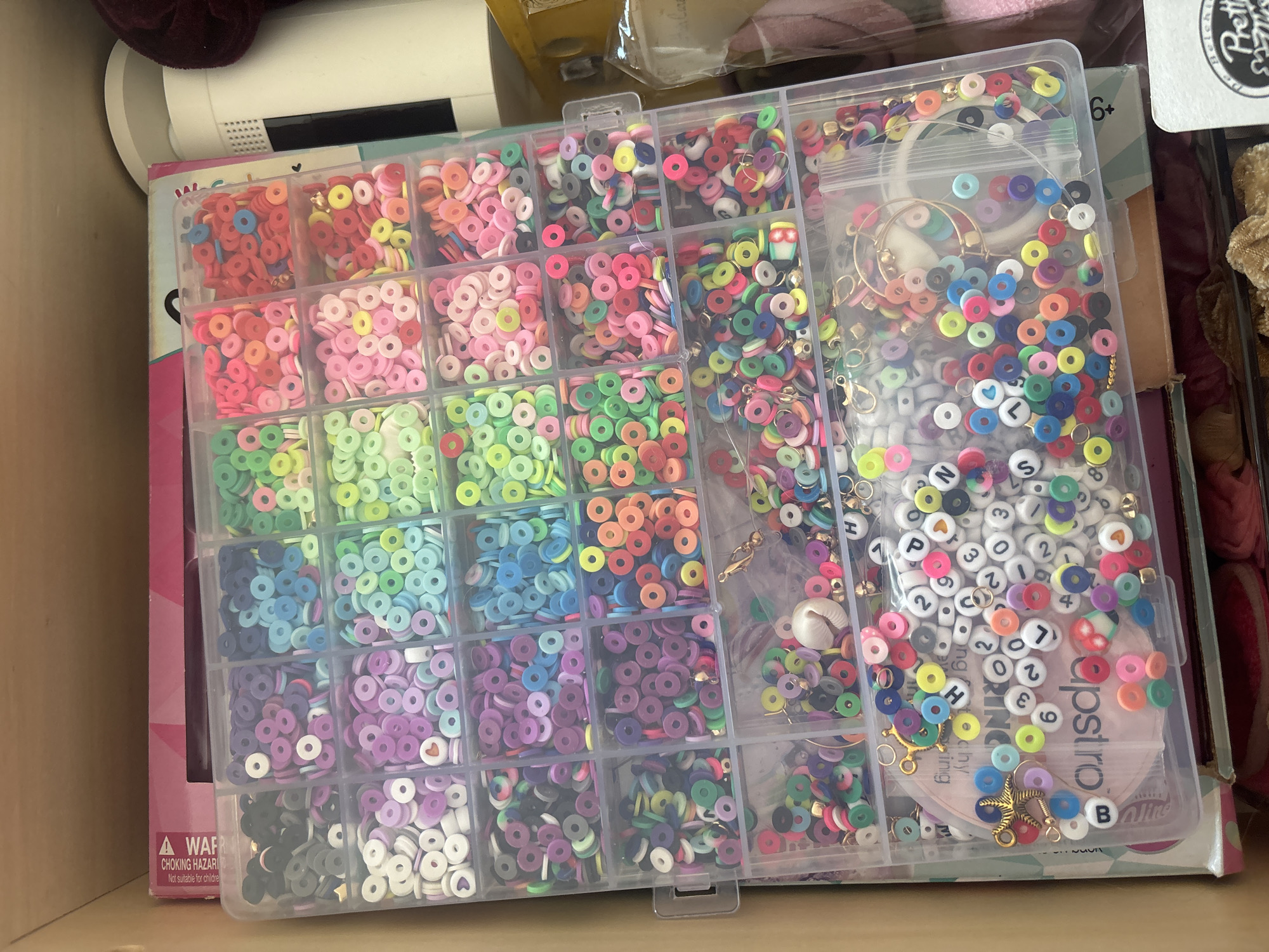 This image depicts a clear plastic container filled with various types of beads, organized into multiple compartments. The beads are predominantly small, flat, circular polymer clay pieces in a wide spectrum of colors, including pinks, blues, greens, yellows, reds, purples, and black. Some compartments also contain letter beads, a few charms (such as a starfish and a seashell), and some metallic findings. The container is resting on top of a pink and white box, which is in turn placed within a cardboard box. The background suggests an indoor setting, likely a storage area or a craft space. There is no discernible information in the image that would allow for identification of a specific location within Dubai, United Arab Emirates. The image is a static, overhead shot of the bead container and its contents.