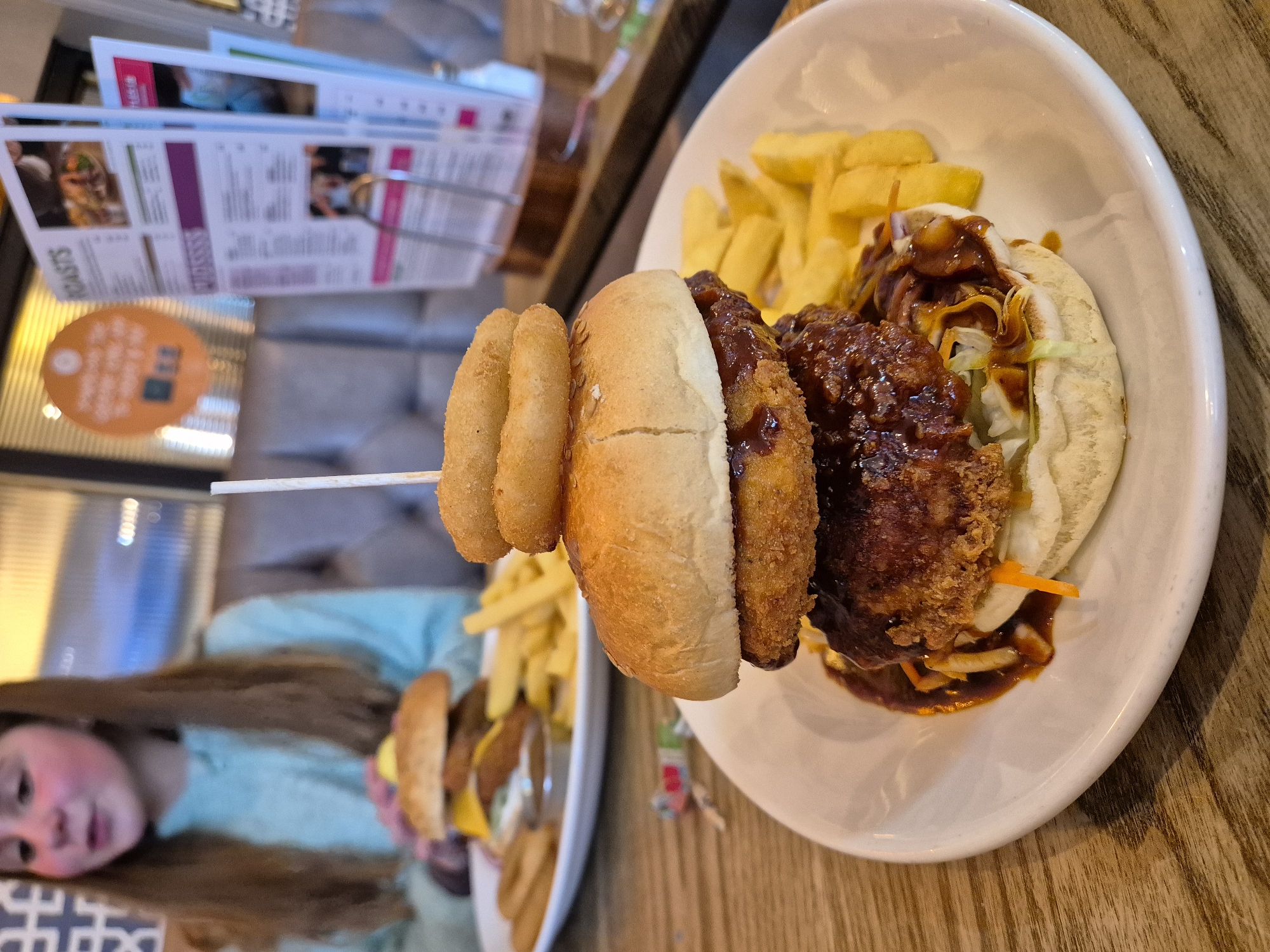 The image shows a plate of food with a burger, fries, and onion rings. The burger appears to have a double patty, and there is a skewer holding the onion rings in place. The food is on a white plate, which sits on a wooden table. In the background, a girl is visible, holding another plate with a similar meal. There is also a menu stand and other dining-related items in the background. The setting seems to be a restaurant in Breckland District, United Kingdom.