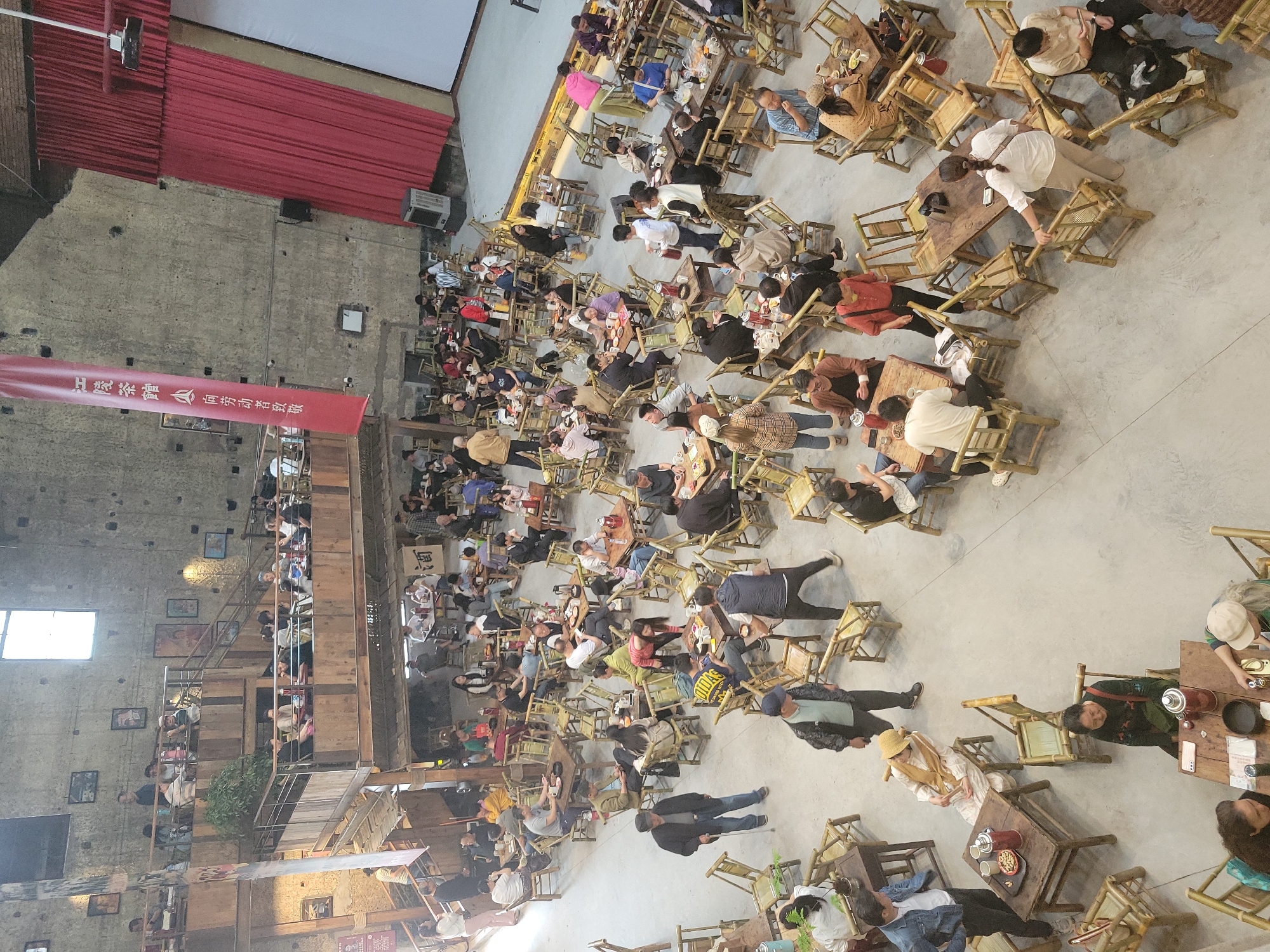 The image shows a high-angle view of a large indoor space populated with people seated at small, square wooden tables and chairs. The seating arrangement is dense and informal. Most people are seated, seemingly engaged in conversation and activities such as eating and drinking. Many people appear to be drinking tea.
The space appears to be a large hall or courtyard with a weathered, industrial feel, suggesting it may be a renovated industrial building. The walls are exposed concrete and brick, and there is a multi-tiered wooden structure along one side, possibly a balcony or mezzanine, also populated with people. A red banner hangs from the structure and has Chinese characters written on it. A red curtain hangs behind the banner, covering a projection screen.
The setting is a commercial establishment, likely a teahouse or similar venue. Given the prompt's reference to Liangjiang, China, it can be concluded that the location is in Liangjiang, China.